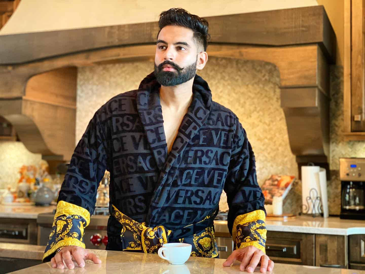 說唱歌手 桌布 壁紙 — 一位留鬍鬚的男人穿著黑色和金色的 Versace 長袍，靠在廚房櫃檯上，手拿咖啡杯。