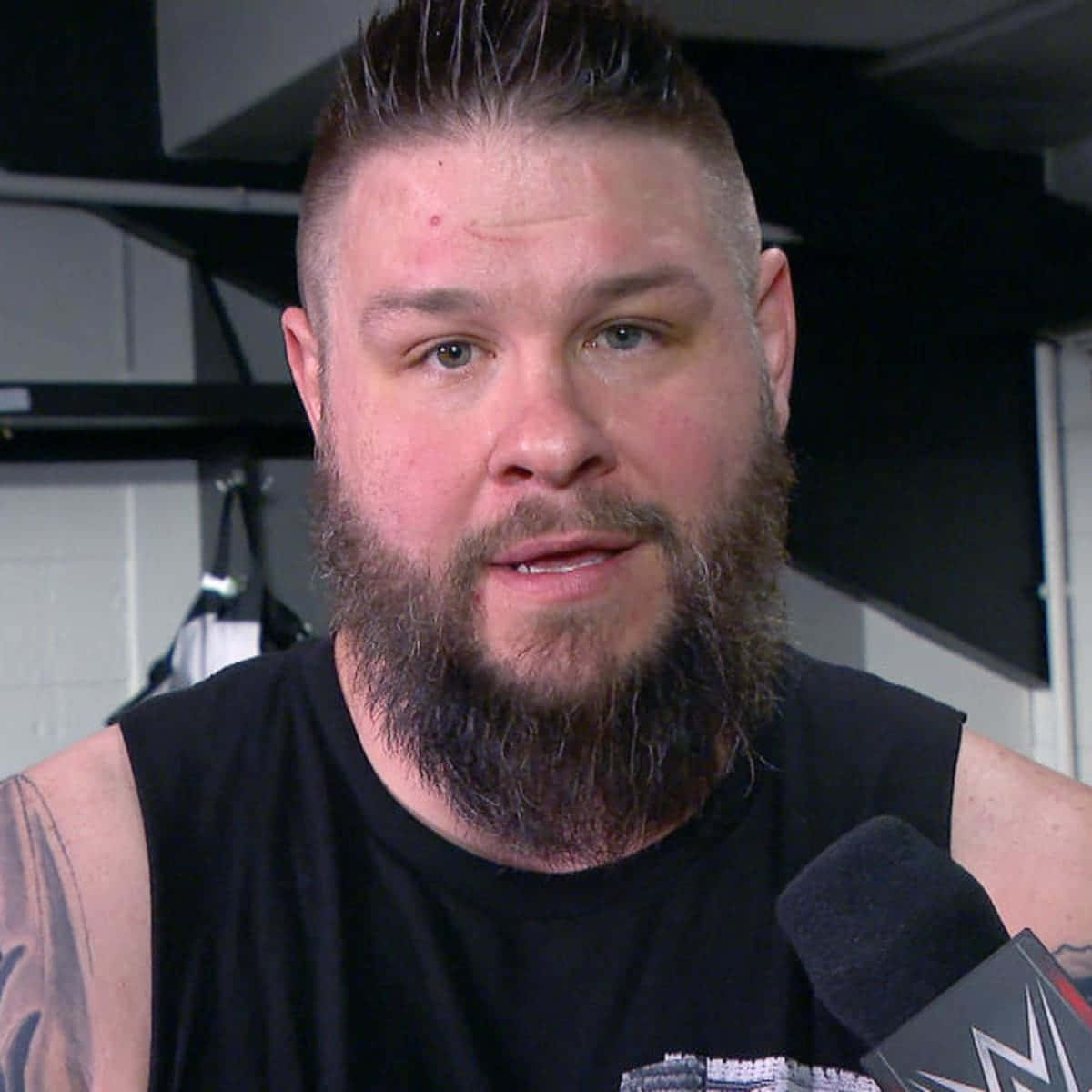 WWE 桌布 壁紙 — 一位留鬍鬚的男人穿著黑色背心，在後台採訪環境中對著WWE麥克風說話。