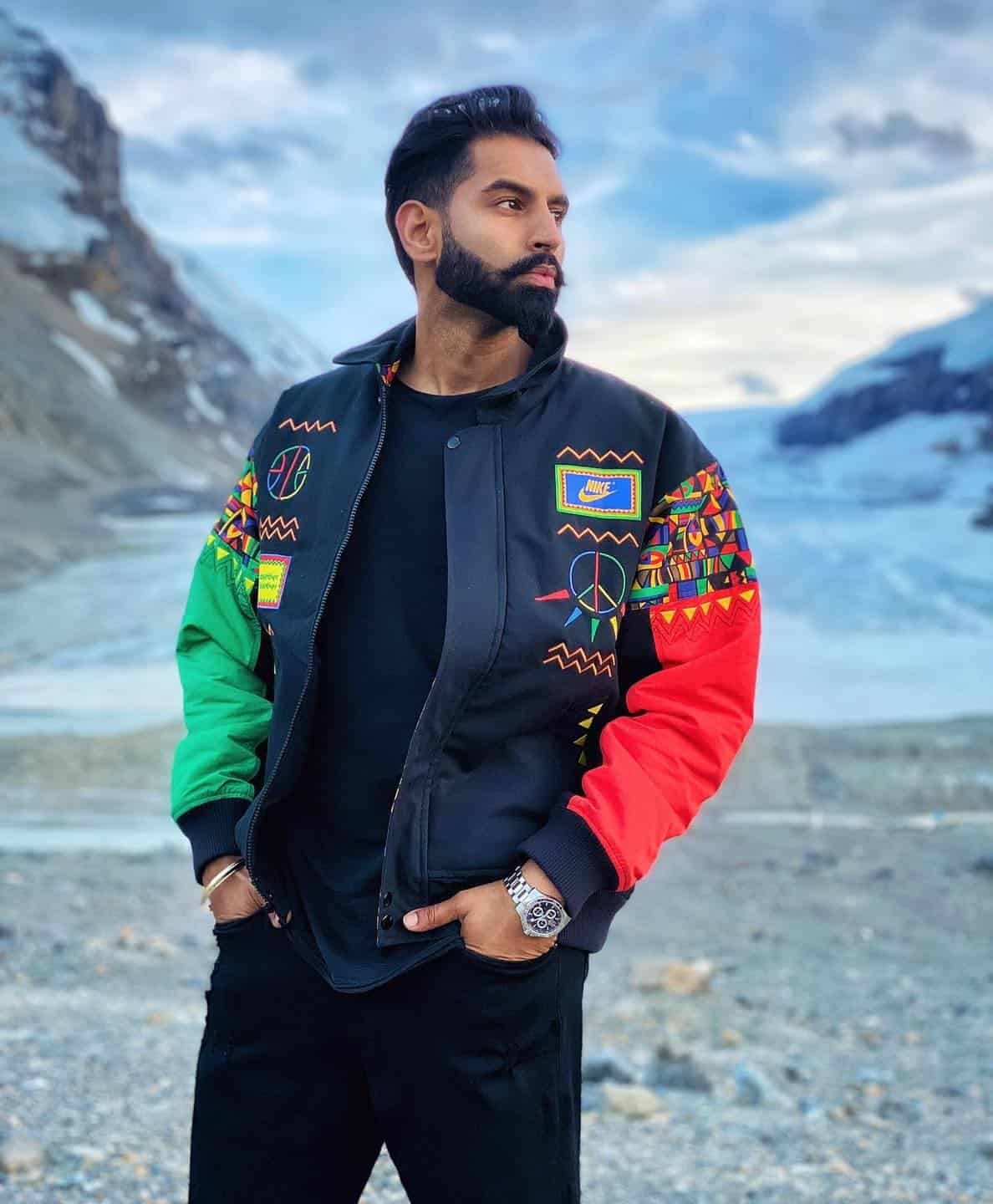 Indian 桌布 壁紙 — 一位留鬍鬚的男人自信地站在雪地山景中，身穿帶有和平符號和幾何圖案的彩色轟炸機夾克。