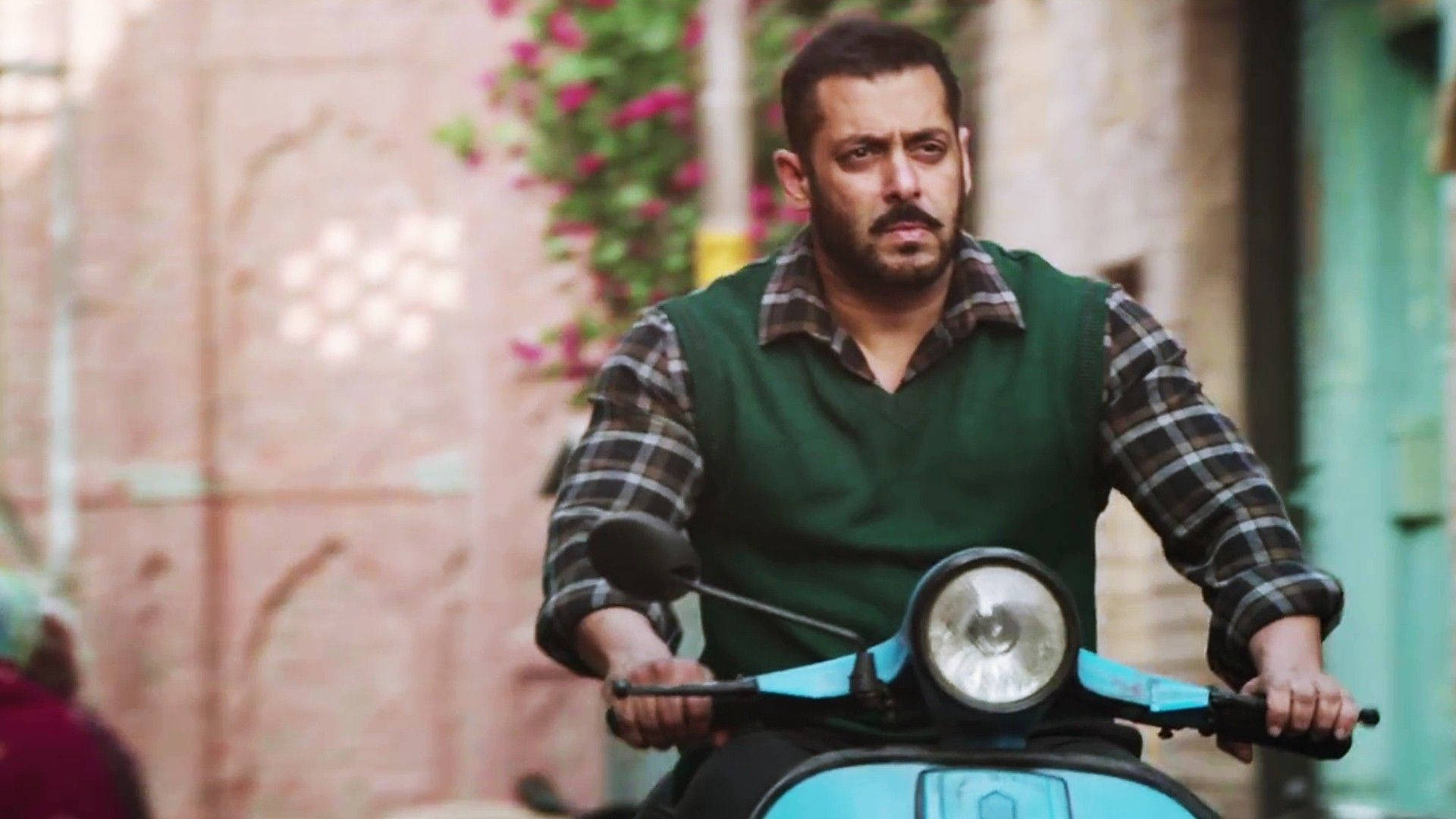 salman khan HD 桌布 壁紙 — 一位留鬍鬚的男人騎著亮藍色滑板車，沿著一條狹窄的小巷行駛，小巷兩旁是粉紅色的牆壁和盛開的藤蔓。