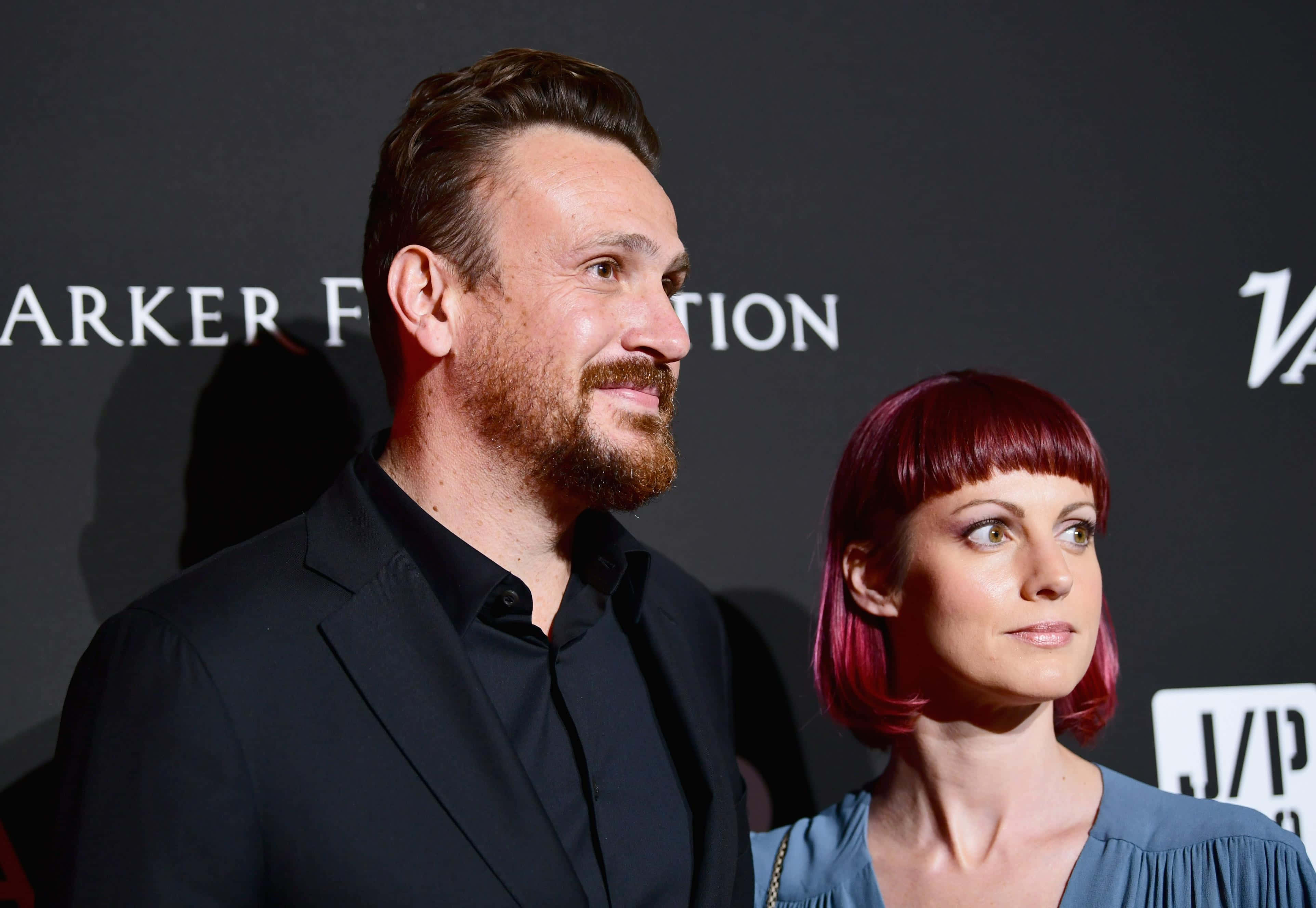 Jason Segel 桌布 壁紙 — 一位留鬍鬚的男士和一位擁有亮麗粉紅色頭髮的女士，在一個正式活動中並肩站立，背景為深色品牌背景。
