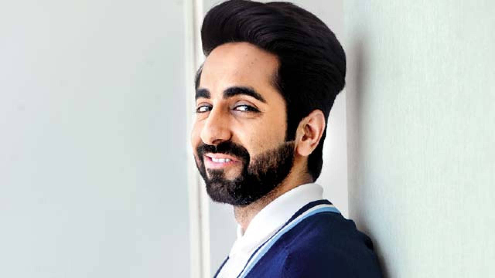 ayushmann khurrana 桌布 壁紙 — 一位留鬍鬚的男士微笑著，輕鬆地靠在一間光線充足的室內環境中的淺色牆上。