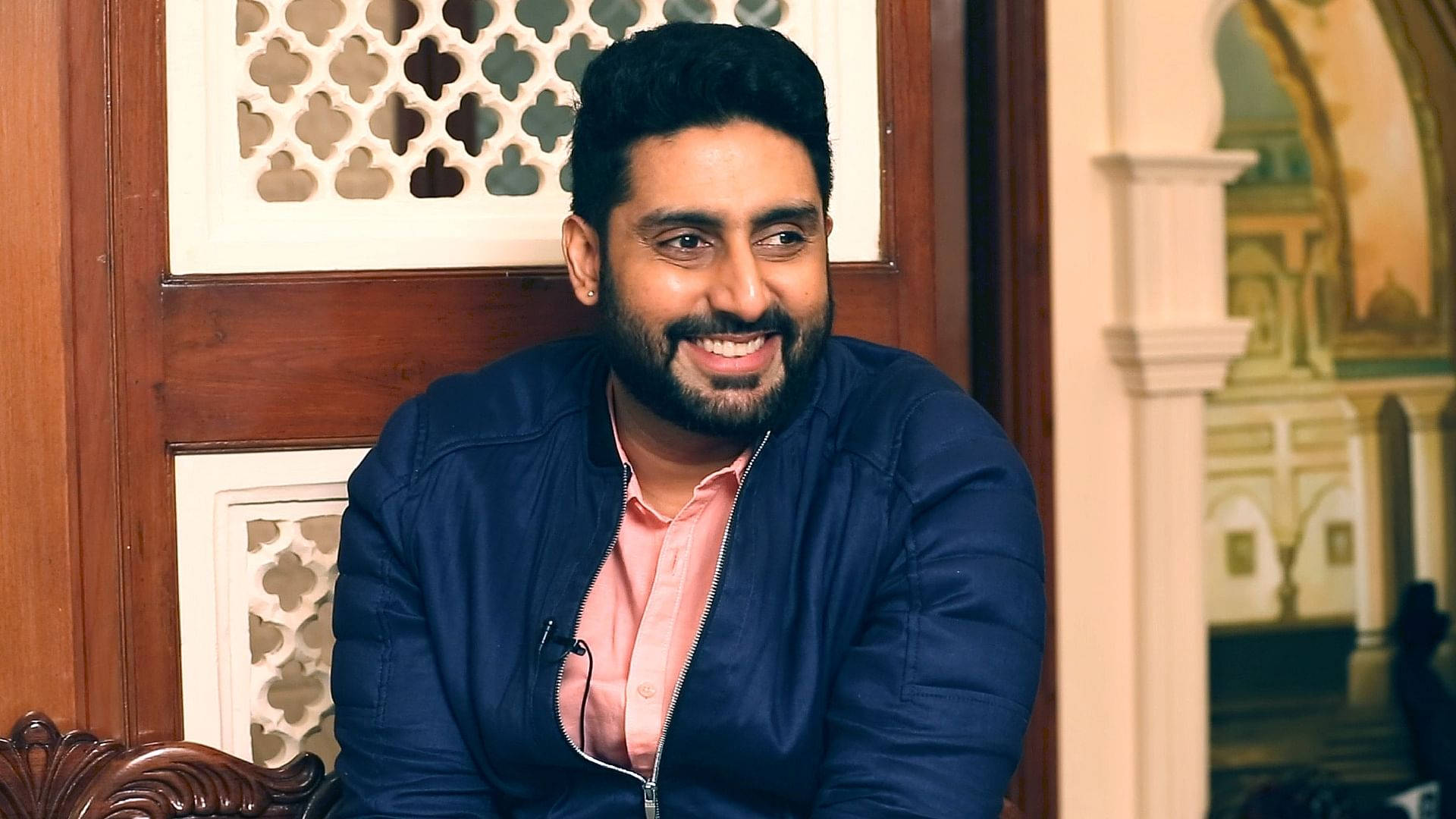 abhishek bachchan 桌布 壁紙 — 一位留鬍鬚的男士微笑著坐在光線溫暖的房間裡，身後有華麗的木製和彩繪建築細節。