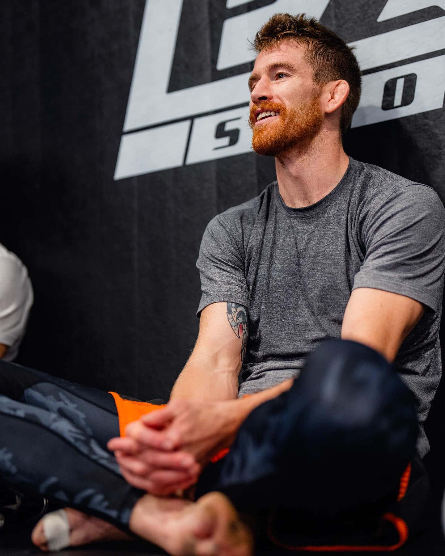 UFC 桌布 壁紙 — 一位留鬍鬚的男士穿著運動服，盤腿坐著微笑，背景帶有品牌標誌。