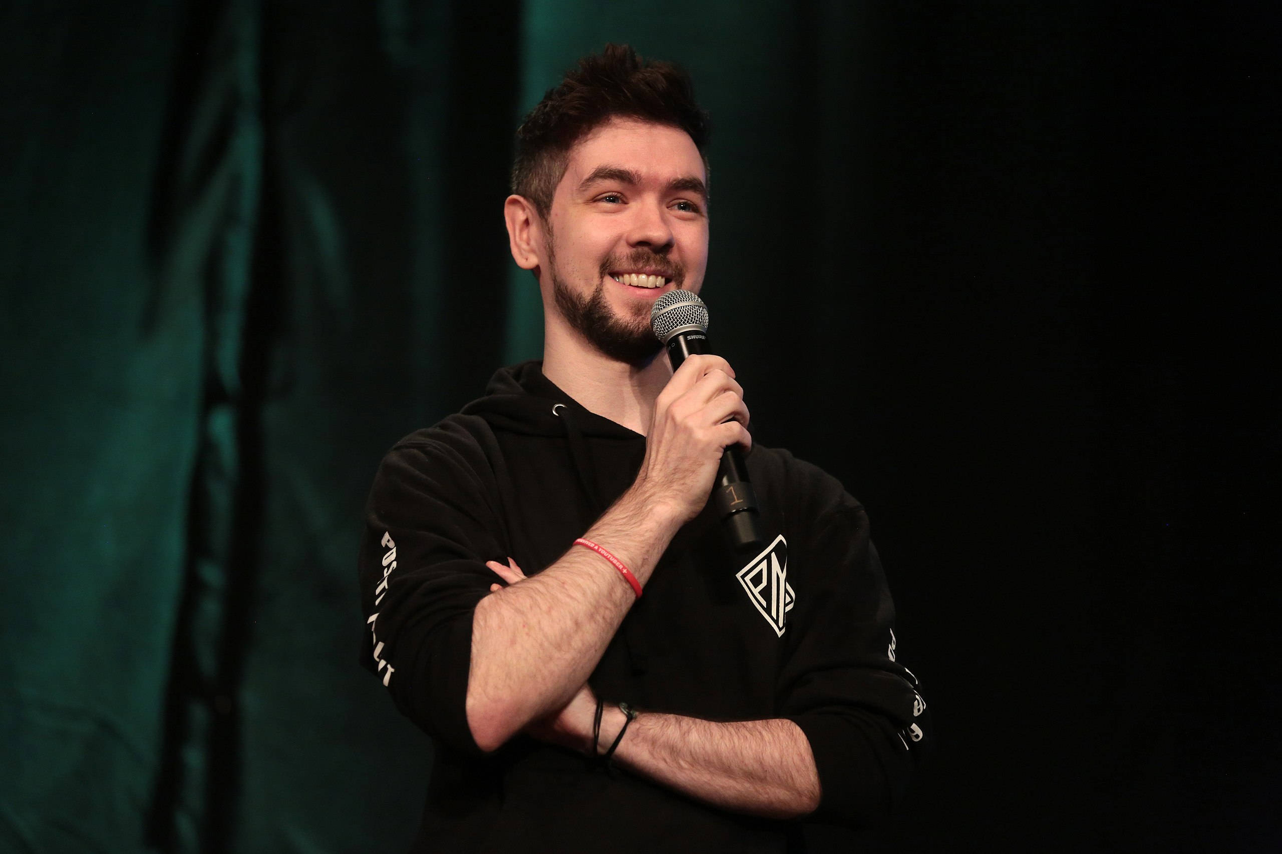 Jacksepticeye 桌布 壁紙 — 一位留鬍鬚的男士面帶微笑，在深綠色窗簾背景下於舞台上手持麥克風。