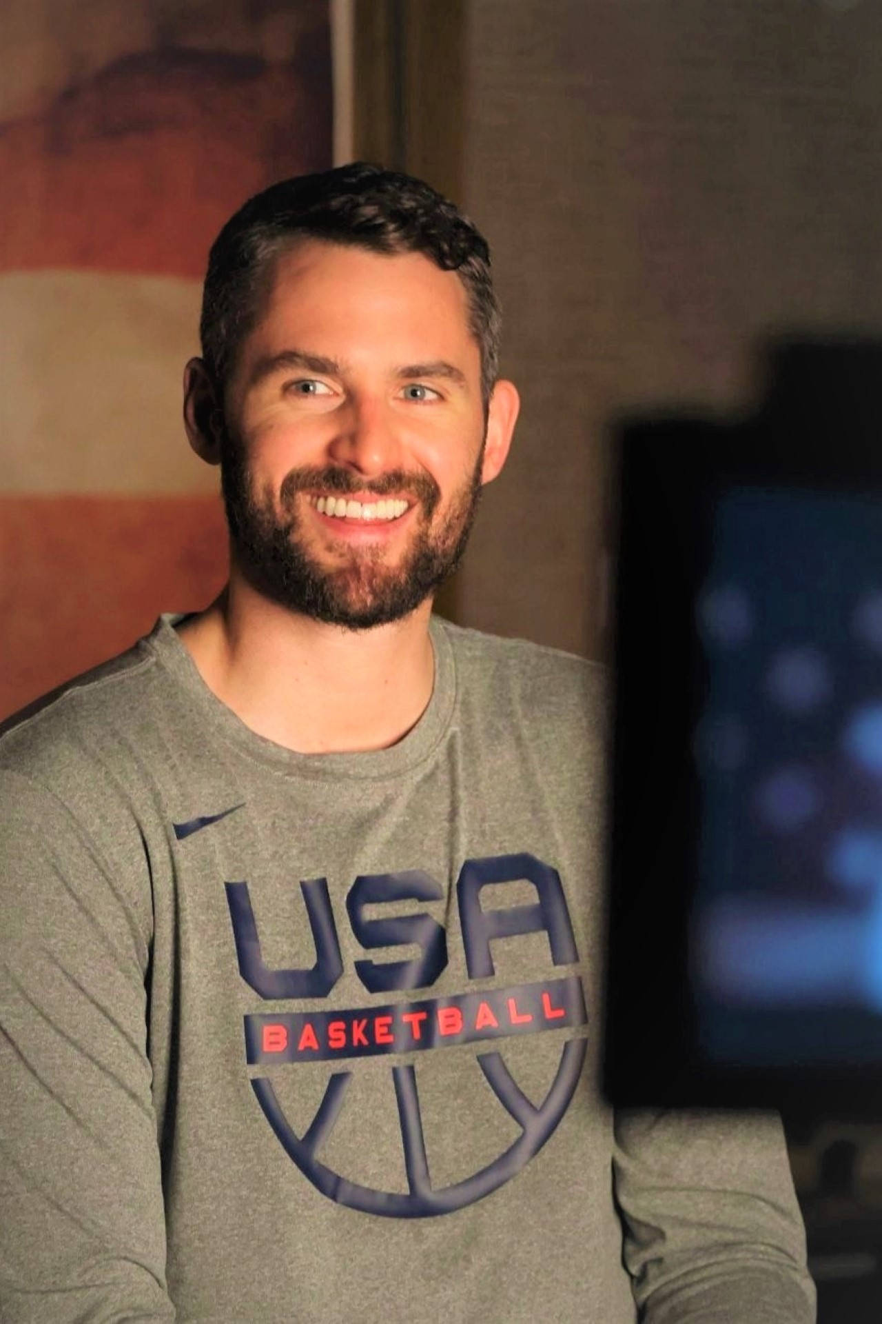 Kevin Love 桌布 壁紙 — 一位留鬍鬚的男士面帶微笑，身穿灰色美國籃球運動衫，在室內以溫暖燈光和模糊背景擺姿勢。