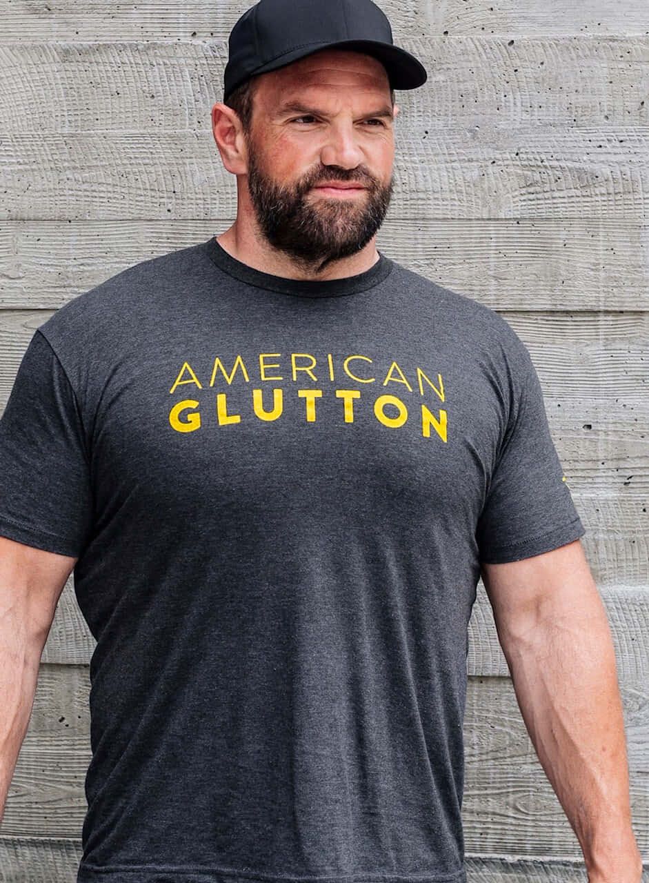 Ethan Suplee 桌布 壁紙 — 一位留鬍鬚的男子，戴著黑色鴨舌帽，身穿深灰色T恤，上面有粗體黃色文字，自信地站在有紋理的混凝土牆前。