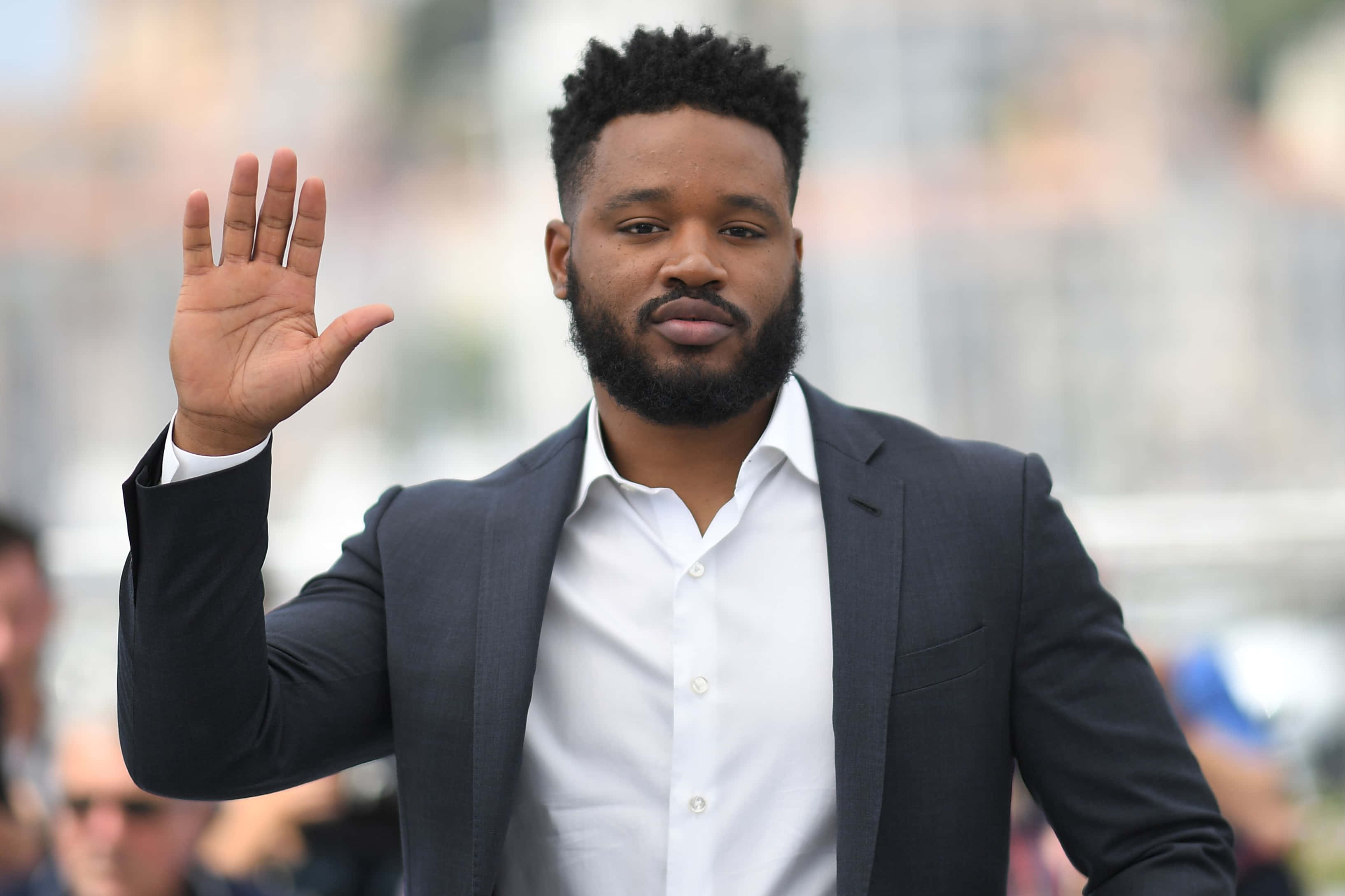 Ryan Coogler 桌布 壁紙 — 一位留鬍鬚的男子，身穿深色西裝和白色襯衫，自信地向鏡頭揮手。