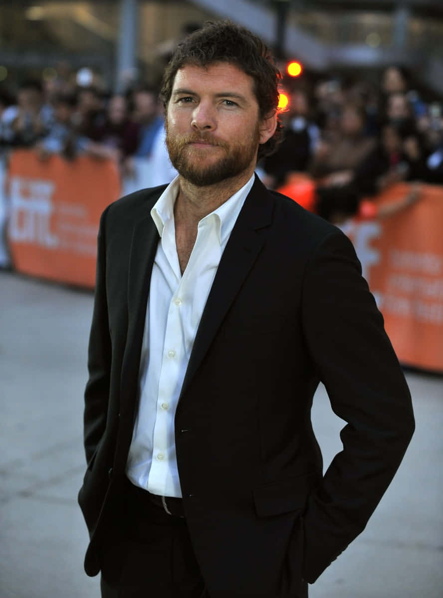 攝影 桌布 壁紙 — 一位留鬍鬚的男子，身穿黑色西裝和白色襯衫，自信地站在紅地毯上，身後是模糊的人群。