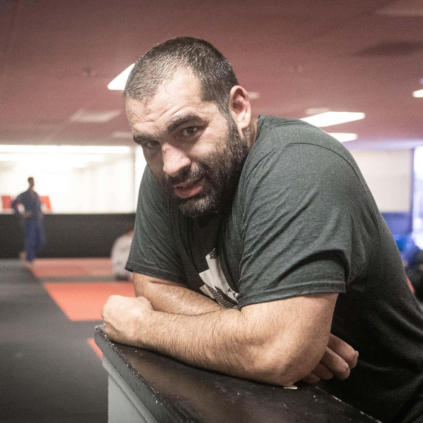 MMA 桌布 壁紙 — 一位留鬍鬚的男子在健身房倚靠在櫃檯上，在明亮的頭頂燈光下帶著專注而強烈的目光。