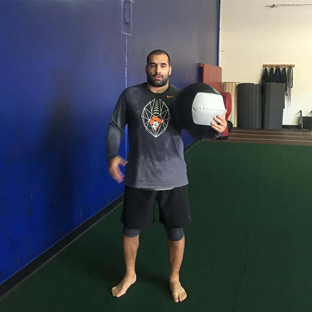 MMA 桌布 壁紙 — 一位留鬍鬚的男子在健身房內，赤腳站在綠色軟墊上，手持藥球，背景是藍色牆壁。