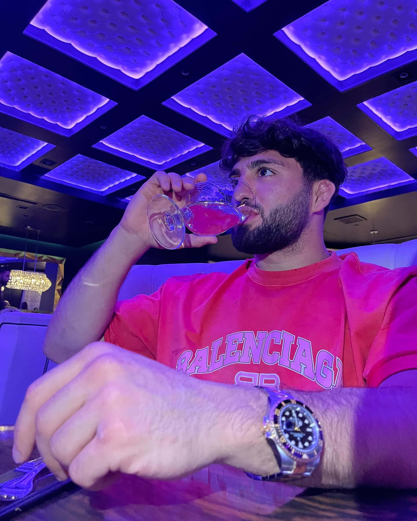 Drinking 桌布 壁紙 — 一位留鬍鬚的男子在昏暗的休息室中，於發光的紫色天花板面板下啜飲粉紅色飲品。