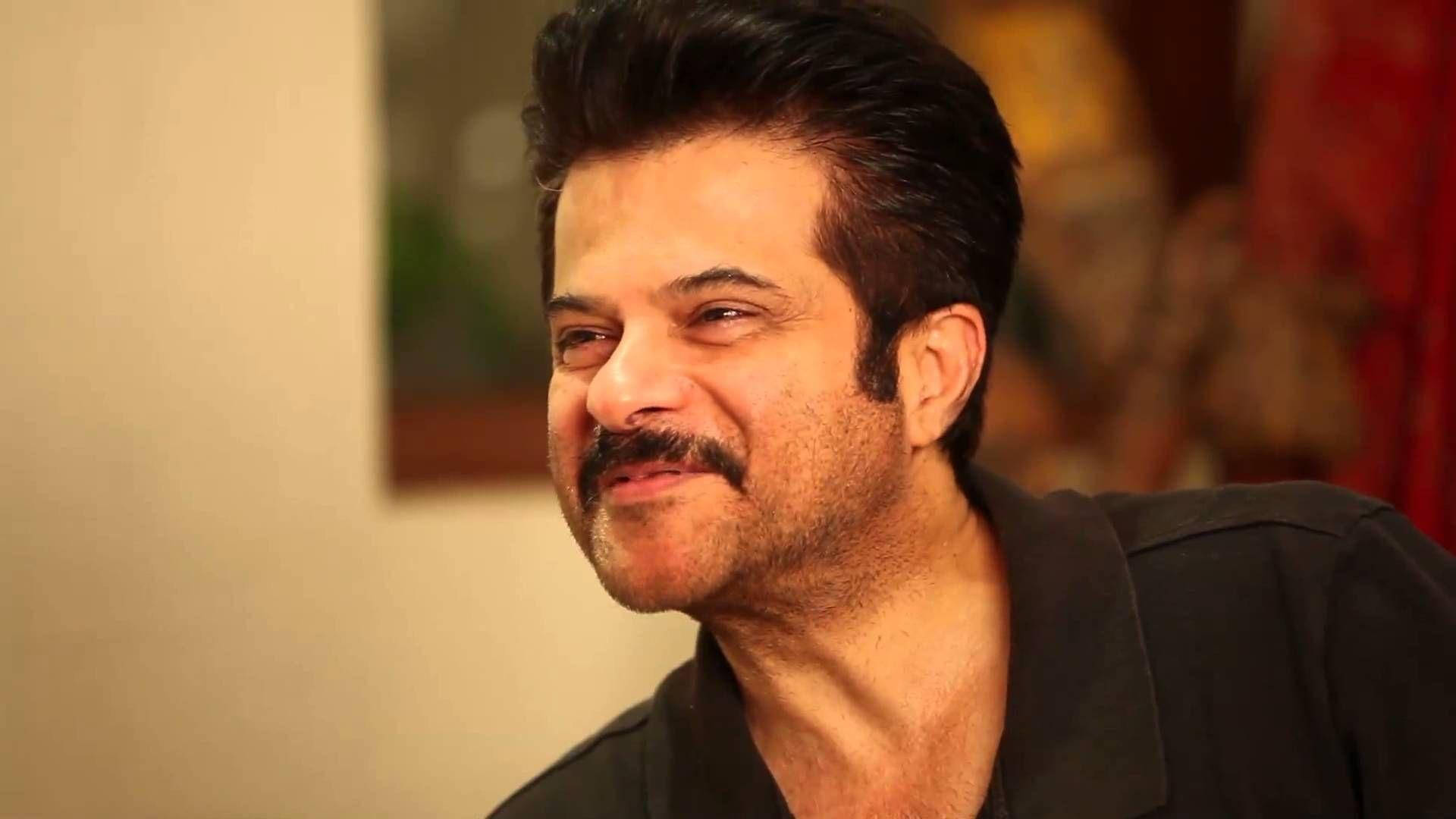 anil kapoor 桌布 壁紙 — 一位留鬍鬚的男子在柔和光線的室內環境中微笑，背景藝術模糊。
