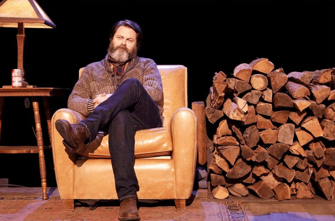 nick offerman 桌布 壁紙 — 一位留鬍鬚的男子在溫暖的舞台燈光下，躺在一張棕褐色皮革扶手椅上，旁邊是一堆堆疊的木柴。