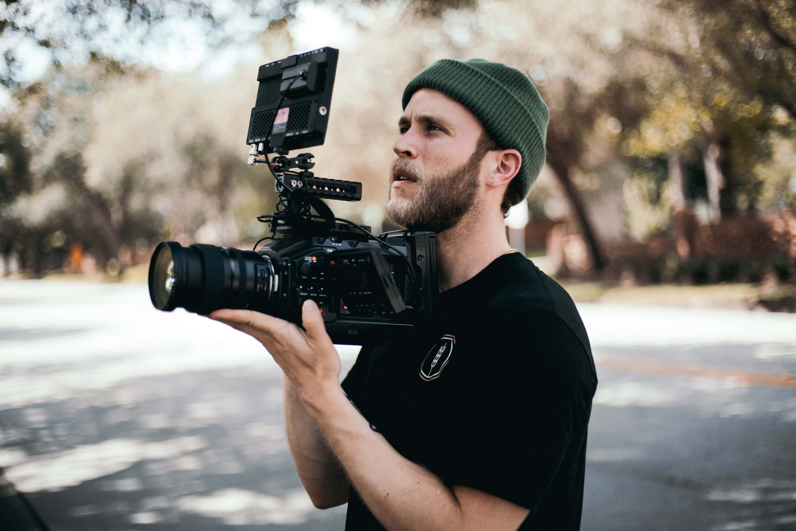 Dslr Camera 桌布 壁紙 — 一位留鬍鬚的男子戴著綠色無簷帽，手持帶有顯示器和鏡頭的專業相機，站在戶外綠樹成蔭的街道上。