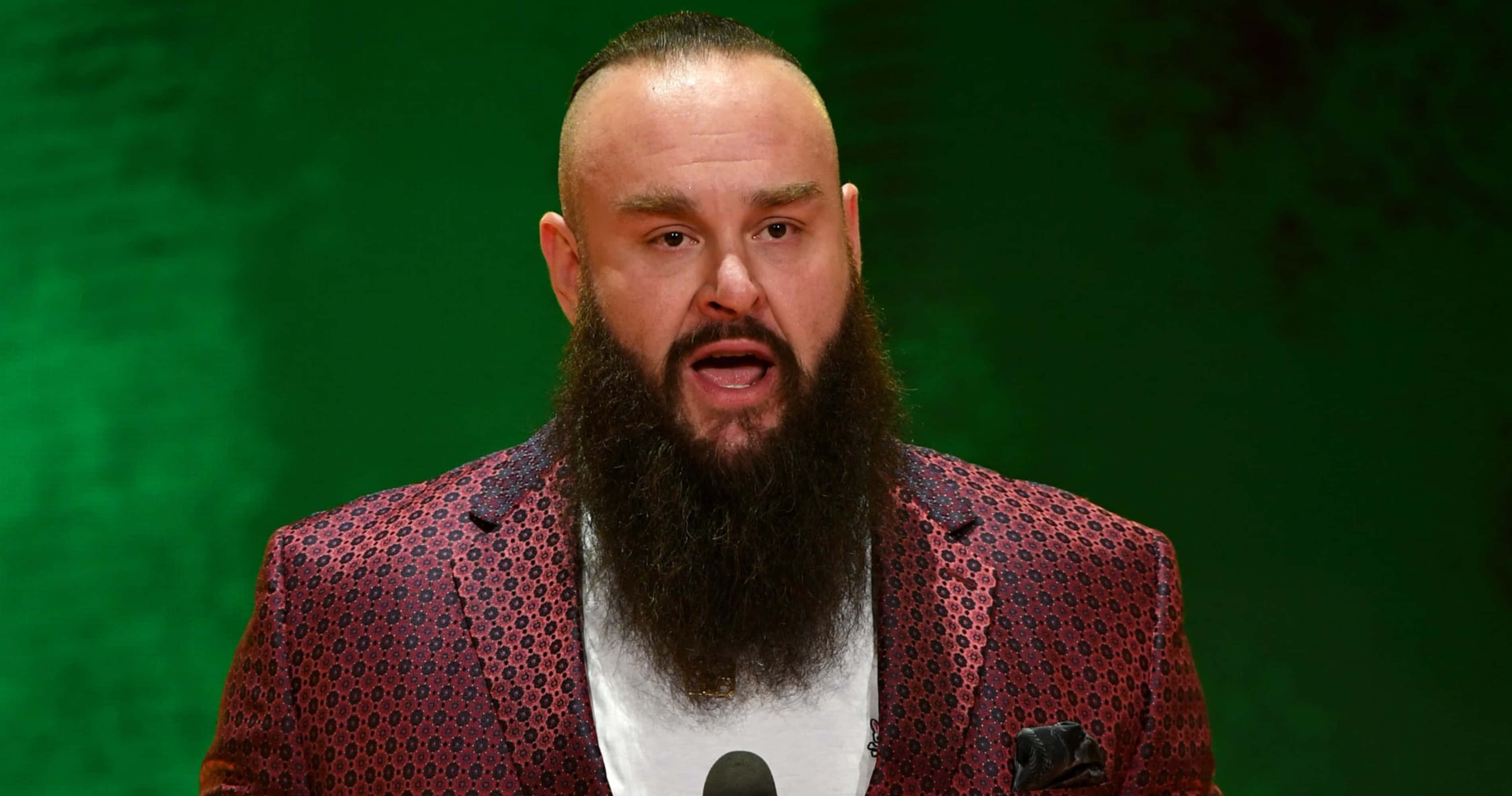 WWE 桌布 壁紙 — 一位留鬍鬚的男子穿著圖案紅色西裝外套，在鮮綠色背景前對著麥克風說話。