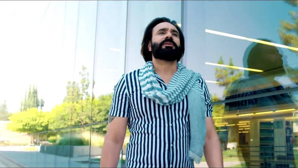 Babbu Maan 桌布 壁紙 — 一位留鬍鬚的男子穿著條紋襯衫和圍巾，自信地站在反射樹木和燈光的玻璃建築前。