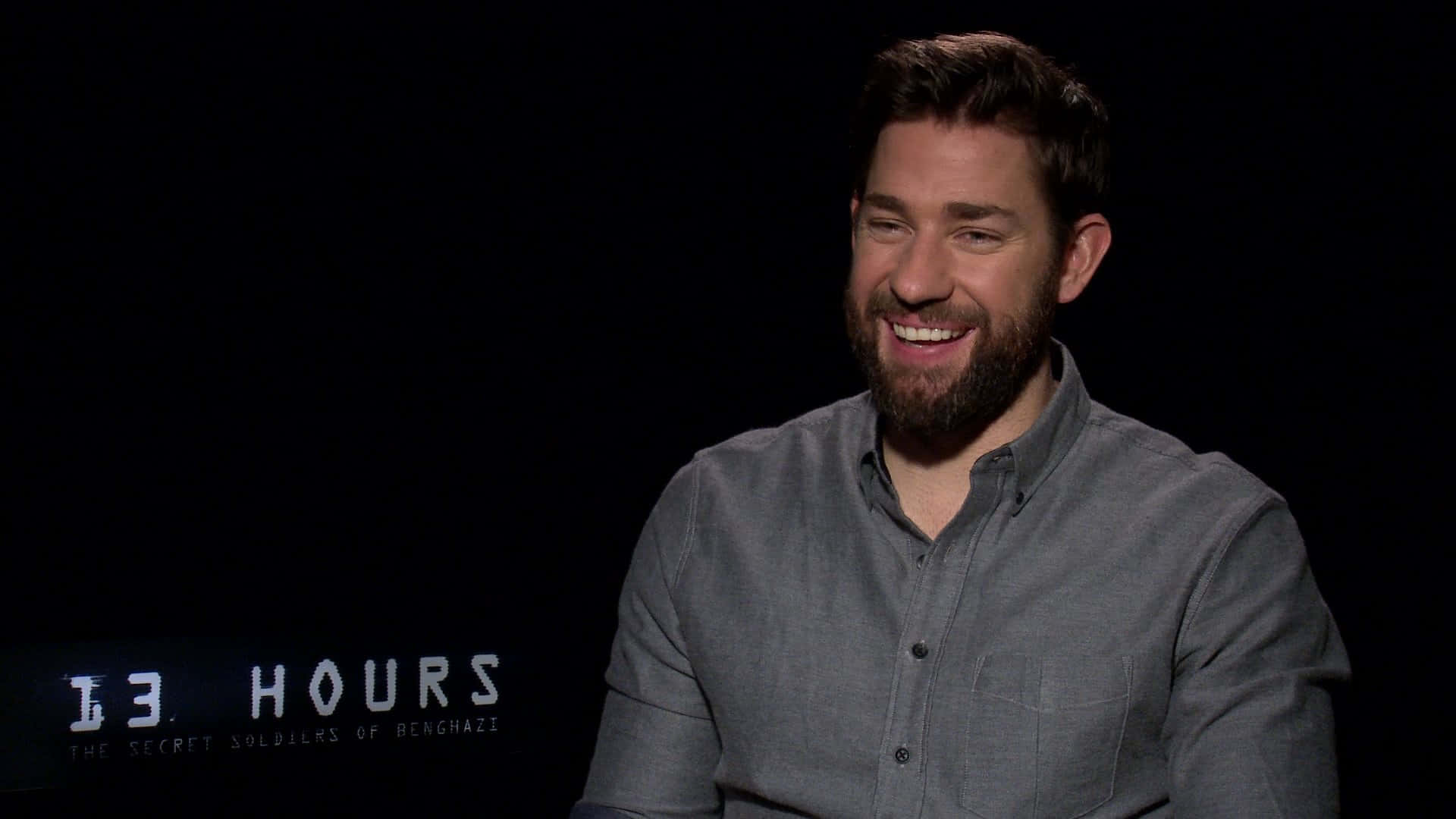 john krasinski 桌布 壁紙 — 一位留鬍鬚的男子穿著灰色襯衫，在黑色背景上帶有電影標題文字的情況下溫暖地笑著。