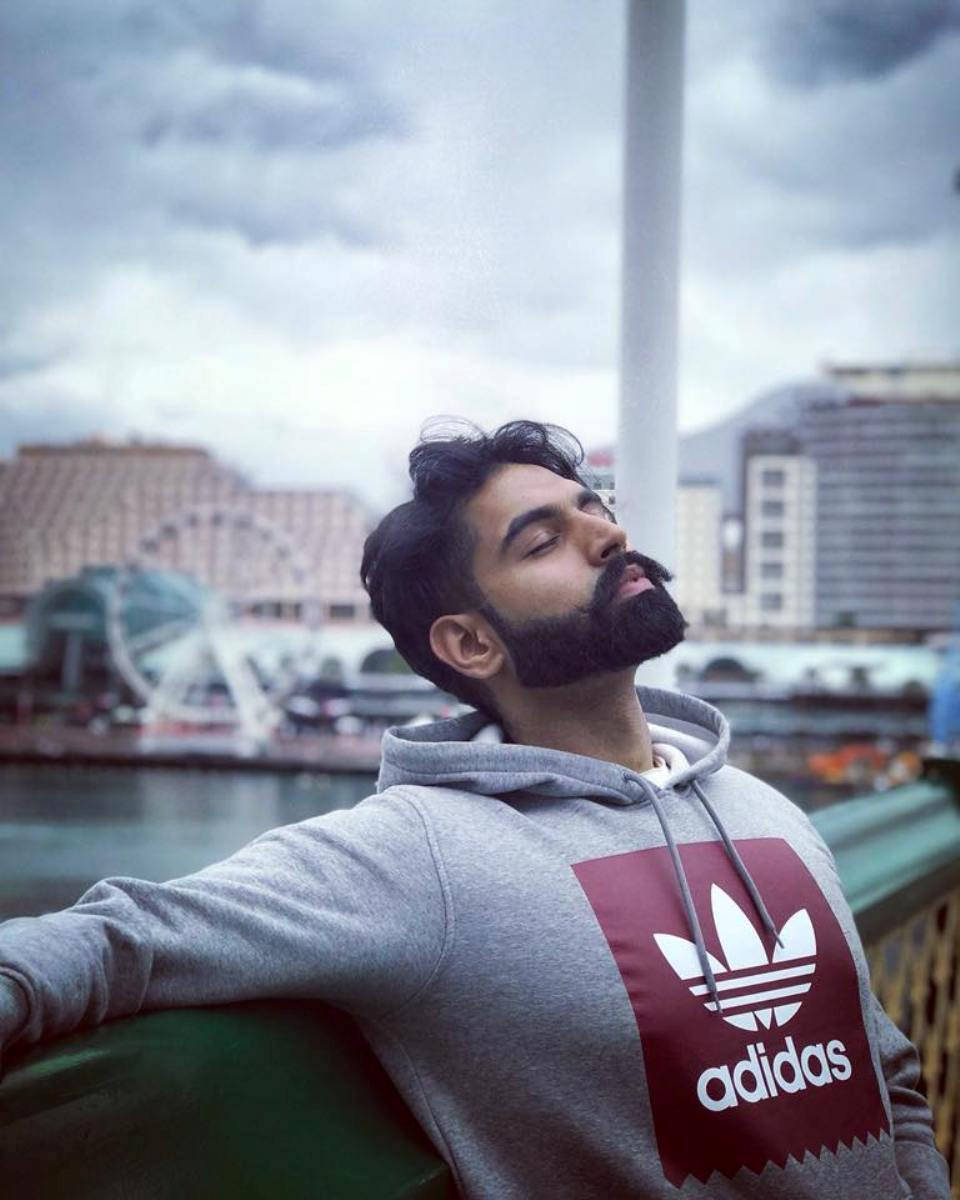 Indian 桌布 壁紙 — 一位留鬍鬚的男子穿著灰色連帽衫，靠在欄杆上，閉著眼睛，享受著多雲天空下霧濛濛的海濱城市景觀。