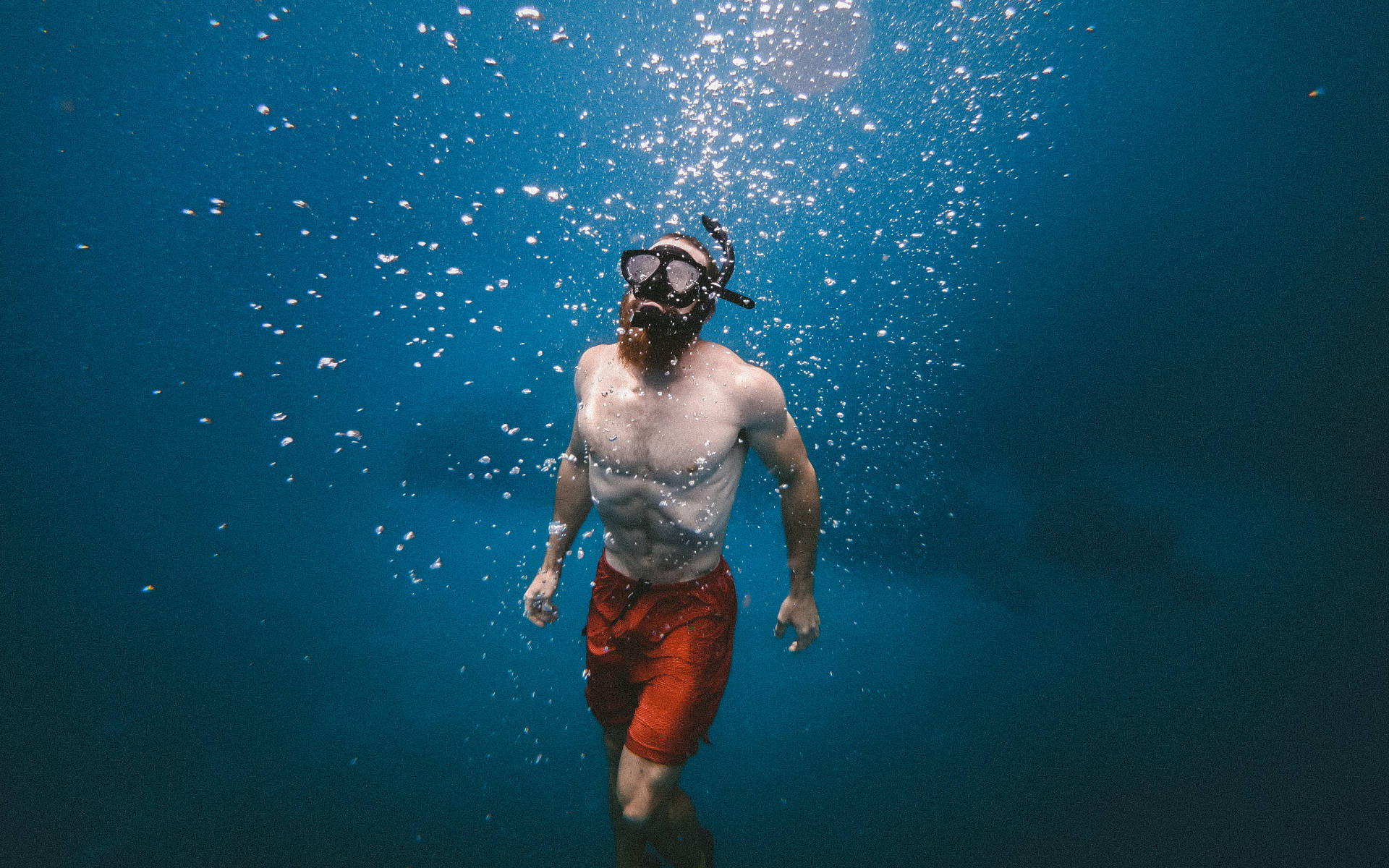 Swimming 桌布 壁紙 — 一位留鬍鬚的男子穿著紅色泳褲和潛水裝備，在清澈的藍色海水中向上游，向陽光照耀的水面釋放氣泡。