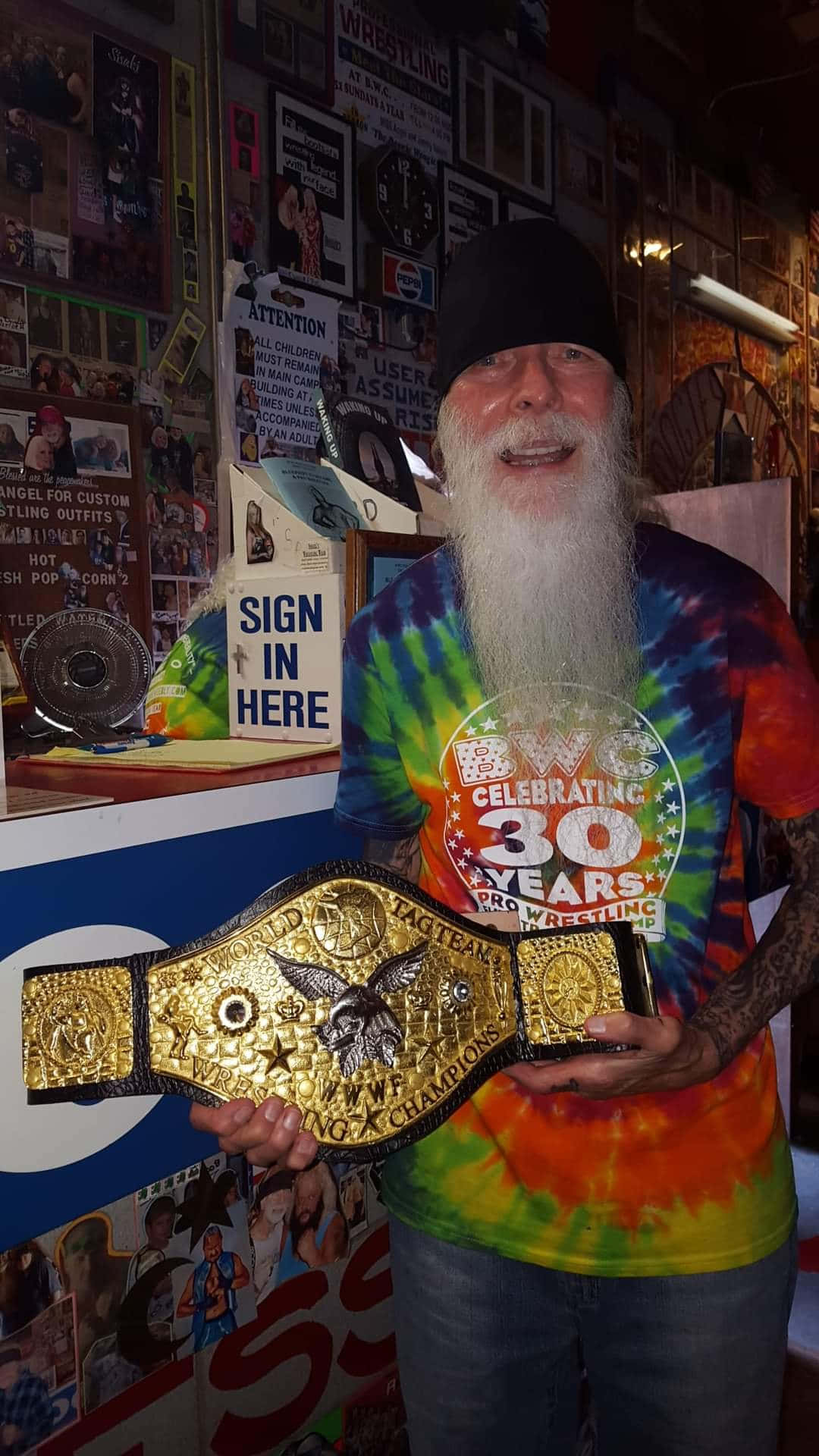 Champion 桌布 壁紙 — 一位留鬍鬚的男子穿著紮染襯衫，在雜亂的職業摔角紀念品房間中自豪地舉起一條金色摔角冠軍腰帶。