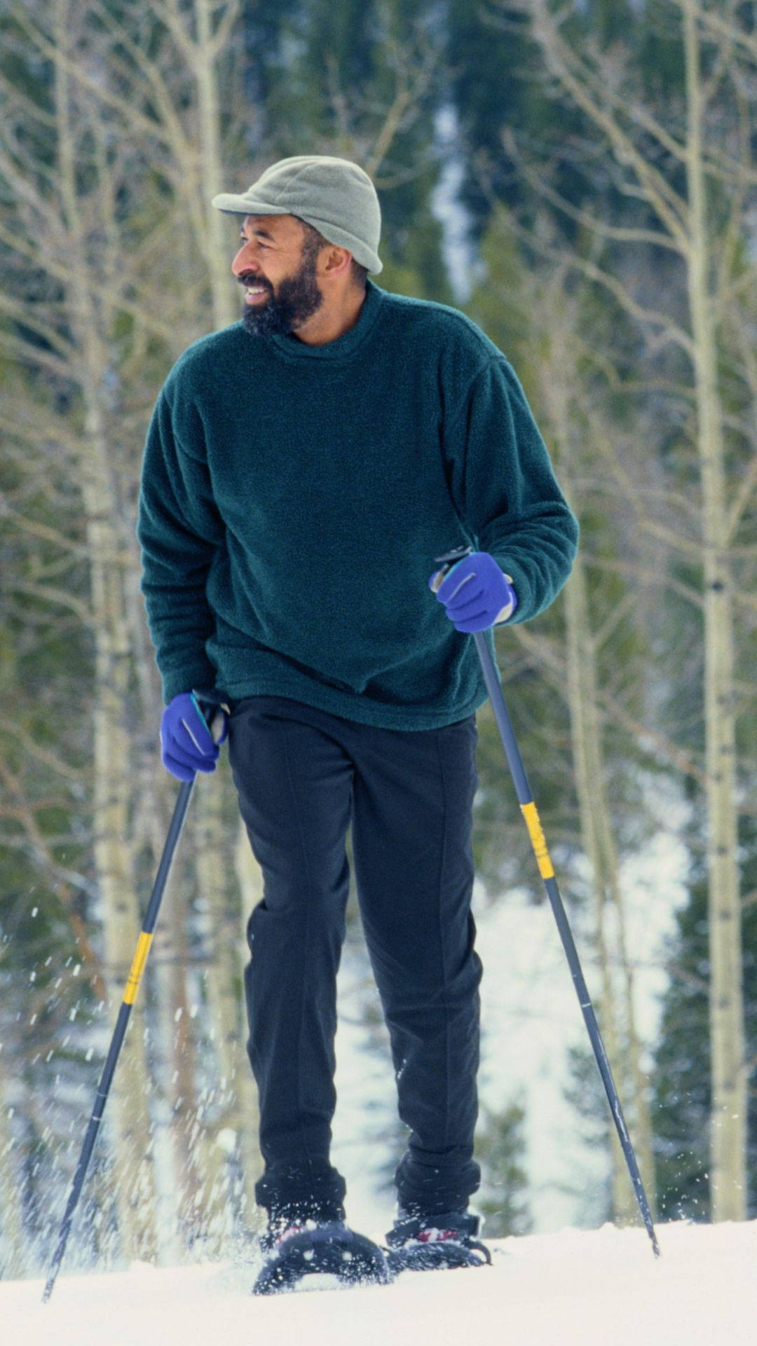 雪 桌布 壁紙 — 一位留鬍鬚的男子穿著綠色毛衣和灰色帽子，在雪地森林中穿著雪鞋行走，微笑著，周圍是光禿禿的樹木。