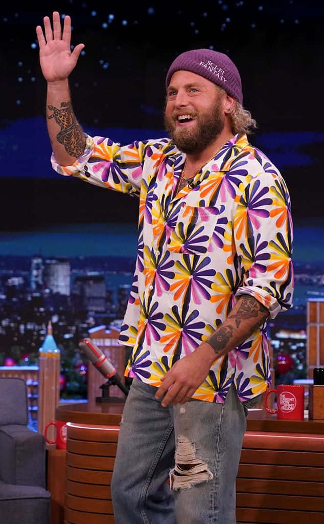 jonah hill 桌布 壁紙 — 一位留鬍鬚的男子穿著色彩繽紛的花襯衫和紫色毛帽，在城市天際線背景的深夜脫口秀舞台上愉快地揮手。