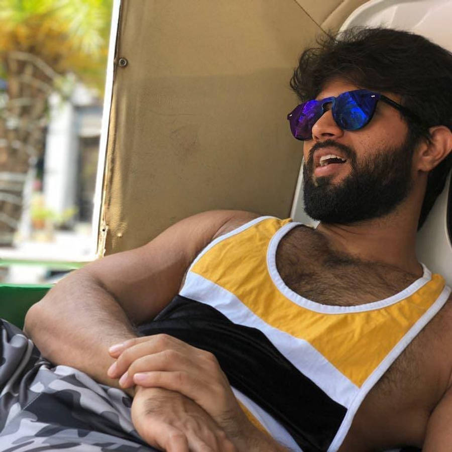 vijay devarakonda 4K 桌布 壁紙 — 一位留鬍鬚的男子穿著黃、黑、白色背心，在嘟嘟車上戴著反光藍色太陽眼鏡休息。