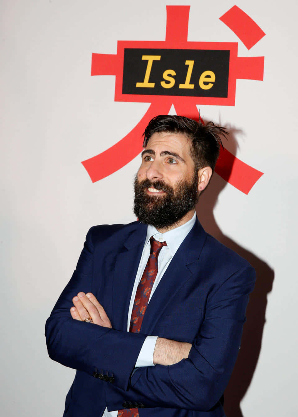 Jason Schwartzman 桌布 壁紙 — 一位留鬍鬚的男子身穿海軍藍西裝，自信地交叉雙臂站在白色牆壁前，牆上有一個大膽的紅色標誌，上面有黃色的「Isle」字樣。