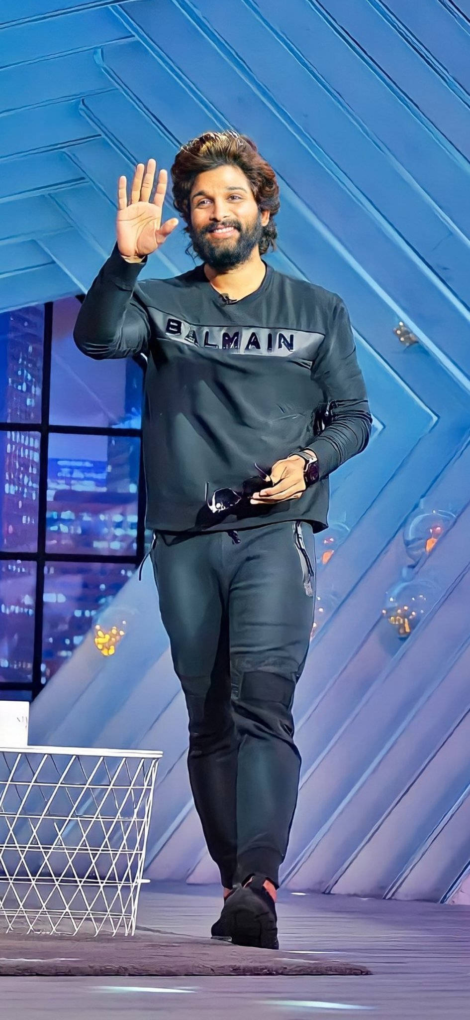 Allu Arjun 桌布 壁紙 — 一位留鬍鬚的男子身穿深色Balmain運動衫，在藍色幾何背景和身後窗戶可見的城市燈光中，邊走邊揮手。