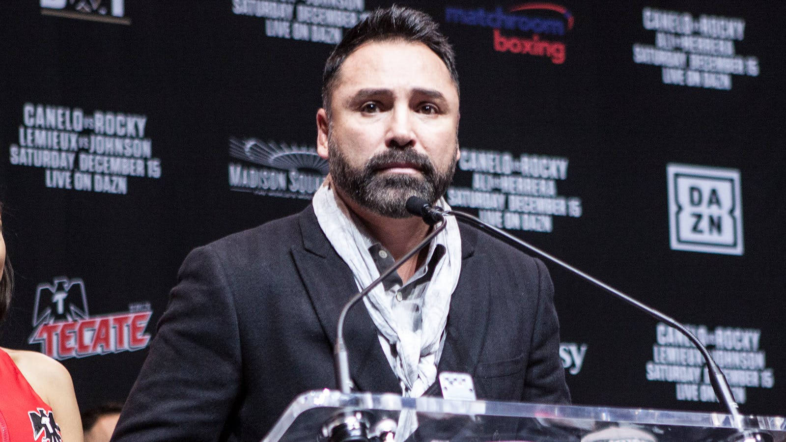 Boxing 桌布 壁紙 — 一位留鬍鬚的男子身穿深色西裝，站在透明講台上，對著麥克風講話，背景為黑色，帶有拳擊活動標誌。