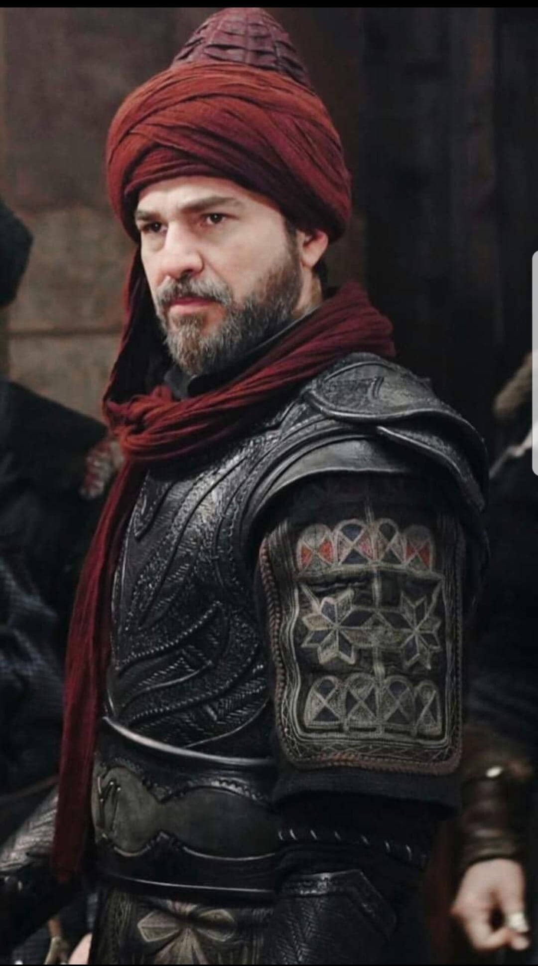 Ertugrul 桌布 壁紙 — 一位留鬍鬚的男子身穿華麗的深色盔甲和紅色頭巾，在昏暗的石頭環境中表情嚴肅地站立。