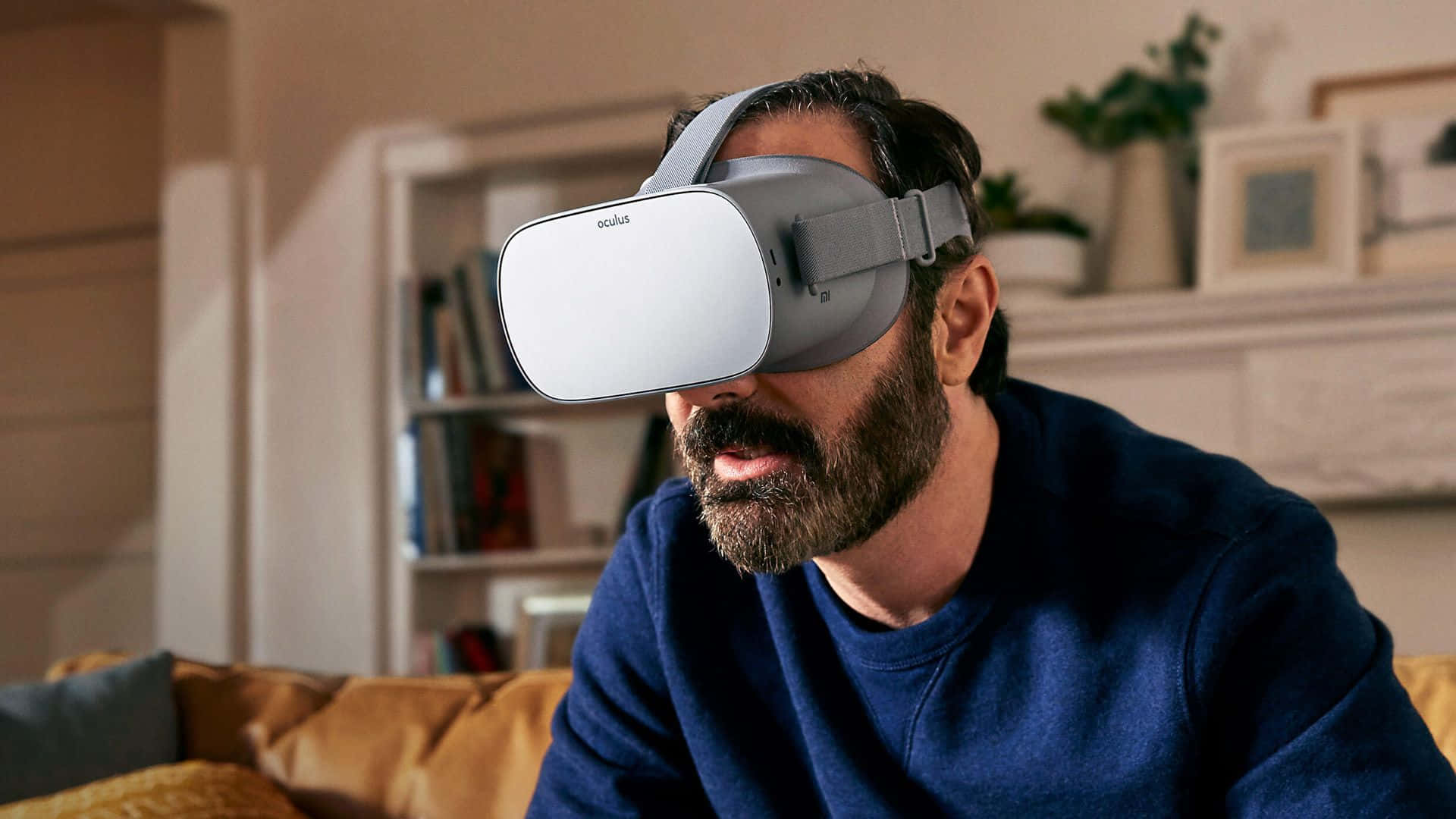 Oculus Quest 2 桌布 壁紙 — 一位留鬍鬚的男子身穿藍色毛衣，坐在光線溫暖的客廳沙發上，戴著白色Oculus VR頭戴式裝置。