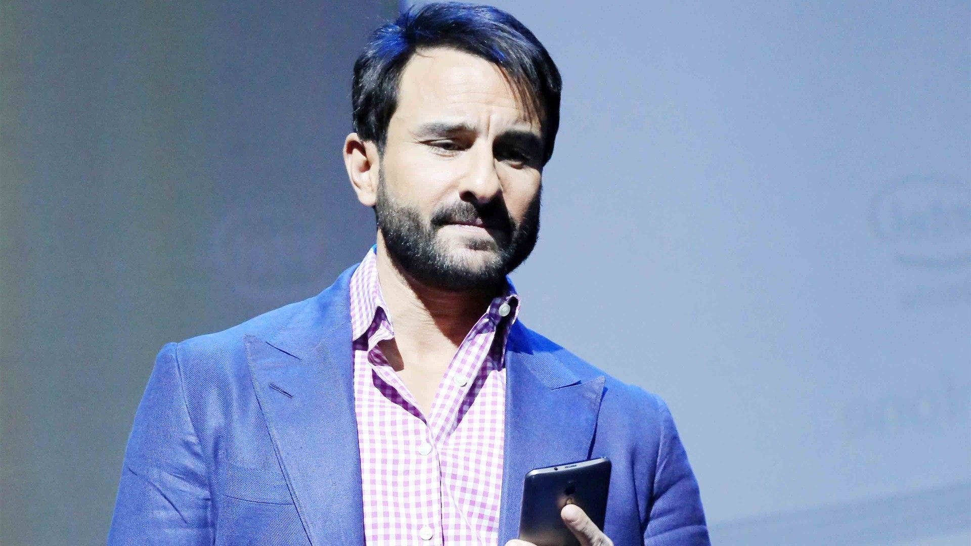 Saif Ali Khan 桌布 壁紙 — 一位留鬍鬚的男子身穿藍色西裝外套和格紋襯衫，站在演講台上手持電話。