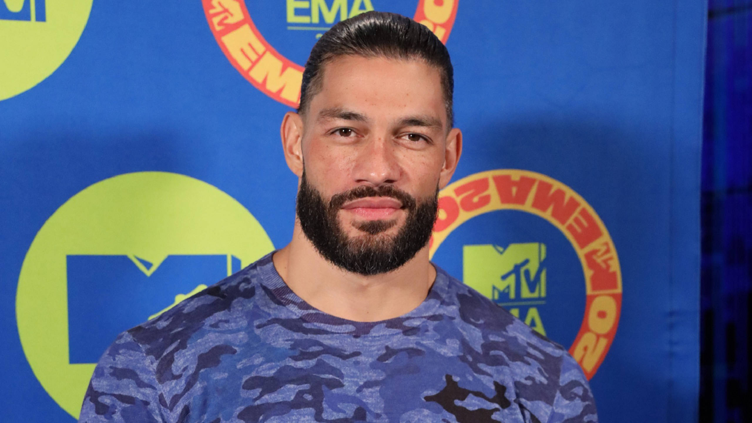 Roman 桌布 壁紙 — 一位留鬍鬚的男子身穿藍色迷彩襯衫，在帶有MTV EMA標誌的鮮豔藍色背景前自信地擺姿勢。