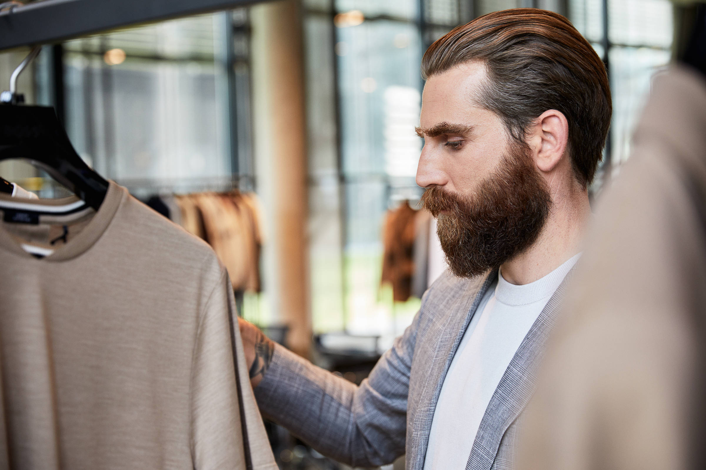 Clothes 桌布 壁紙 — 一位留鬍鬚的男子身穿訂製西裝外套，在現代服裝店仔細檢查掛在衣架上的米色T恤。