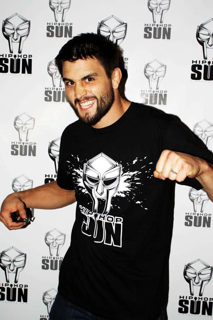 UFC 桌布 壁紙 — 一位留鬍鬚的男子身穿黑色 'Hip Hop Sun' T恤，在品牌活動背景前自信地舉起拳頭擺姿勢。