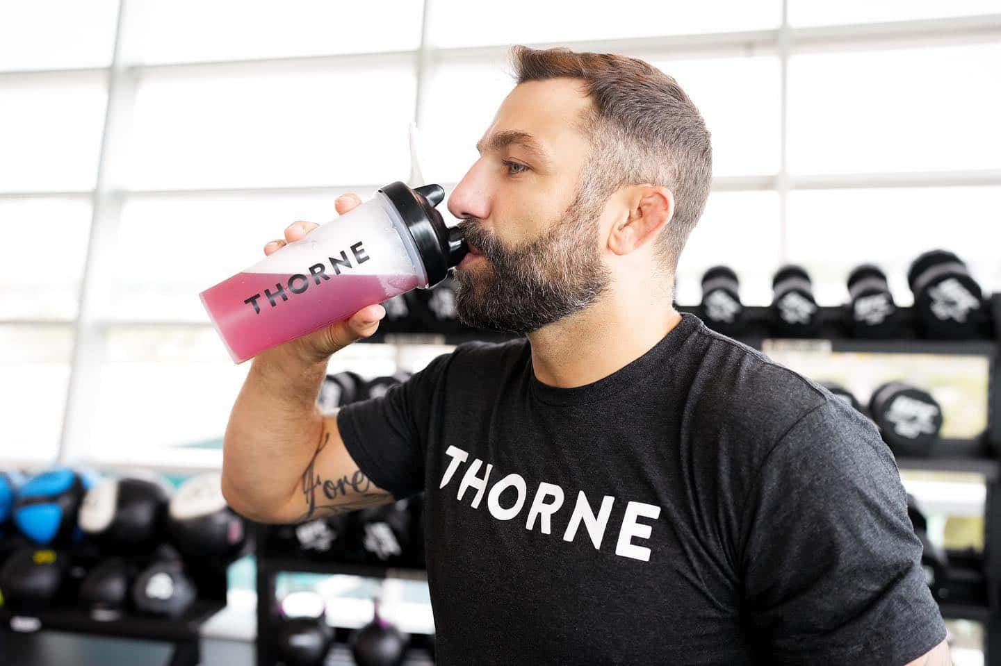 健身房 桌布 壁紙 — 一位留鬍鬚的男子身穿黑色Thorne T恤，在明亮的健身房裡用搖搖杯喝粉紅色奶昔。