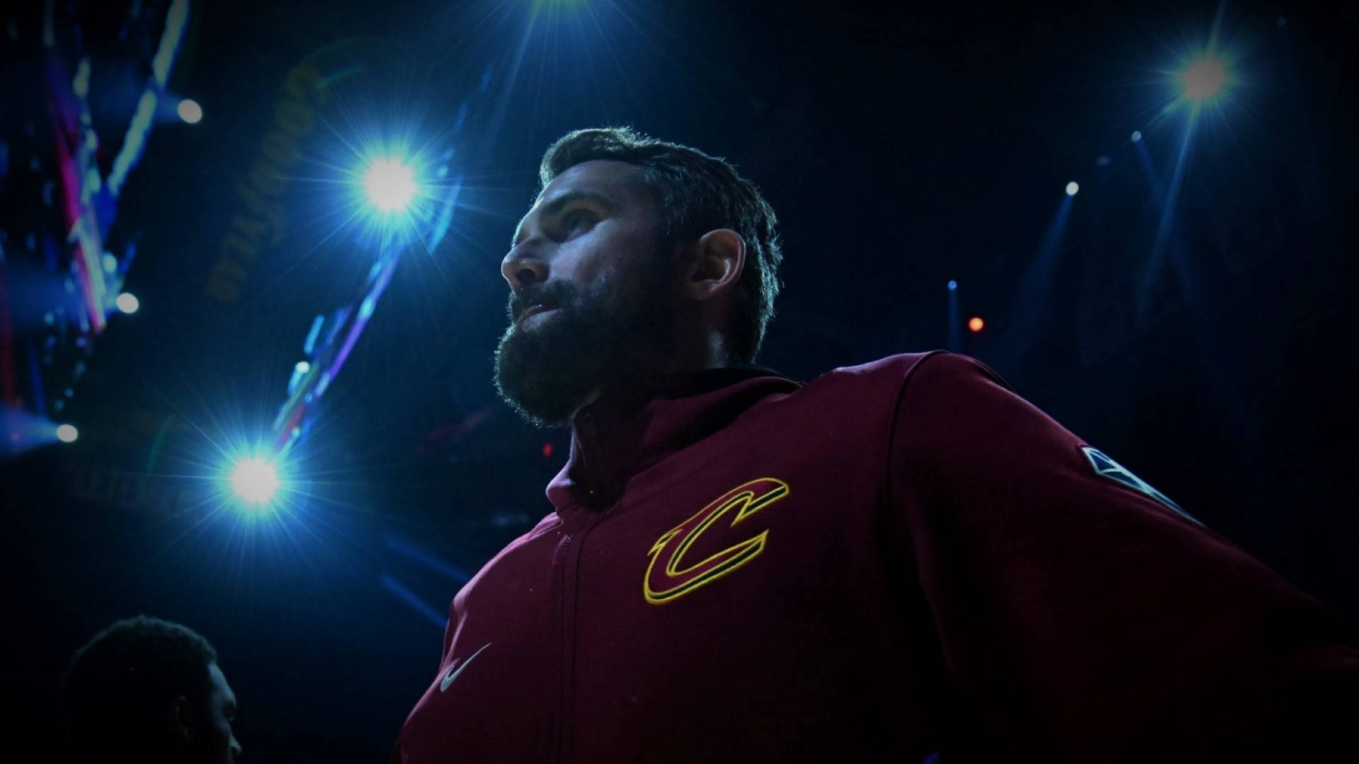 Kevin Love 桌布 壁紙 — 一位留鬍鬚的籃球教練穿著栗色克里夫蘭球衣，在昏暗的場館中站在明亮的聚光燈下。