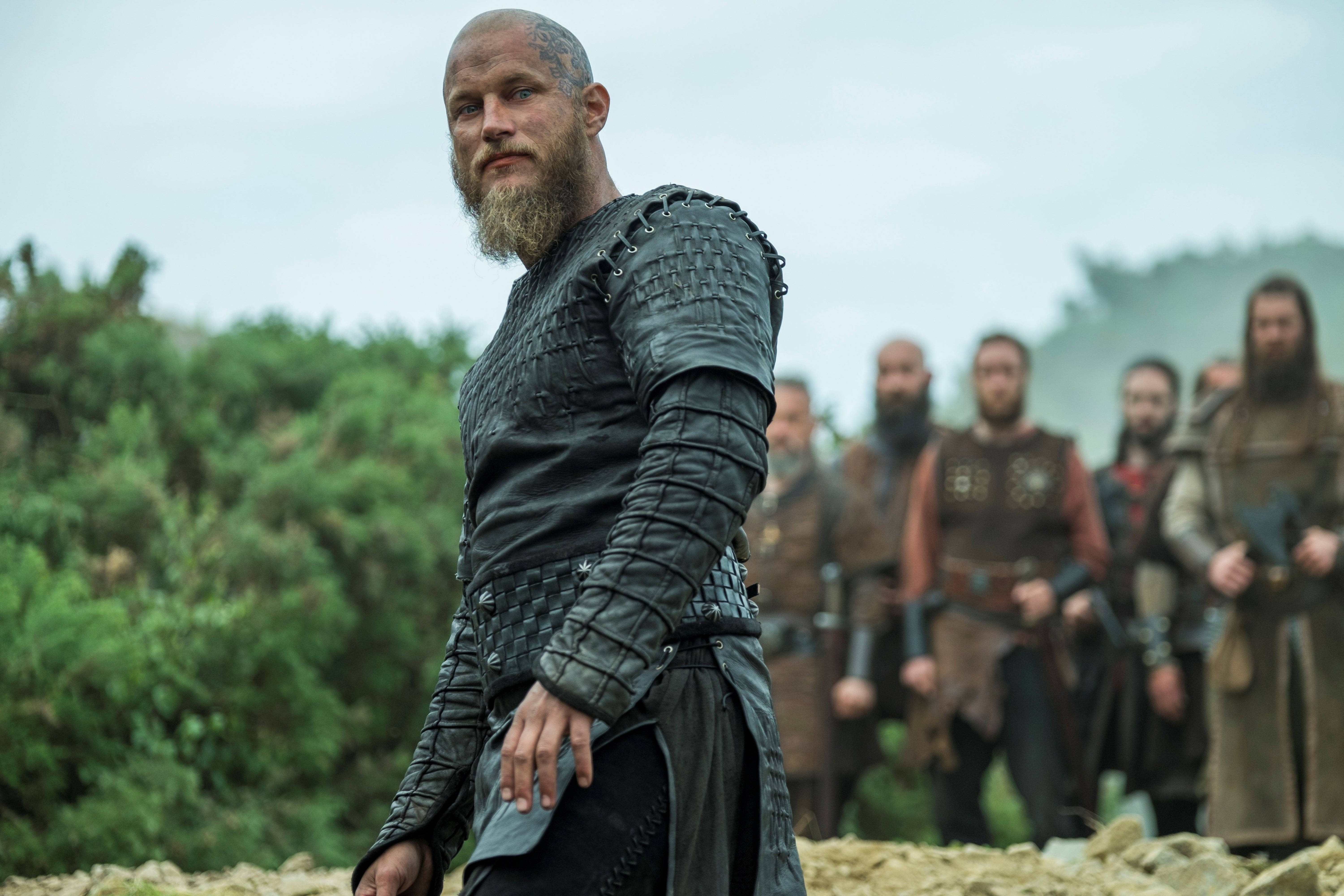 Vikings 桌布 壁紙 — 一位留鬍鬚的維京戰士身穿深色皮革盔甲，在岩石地上與他的部下站在霧氣瀰漫的背景中，顯得堅定不移。