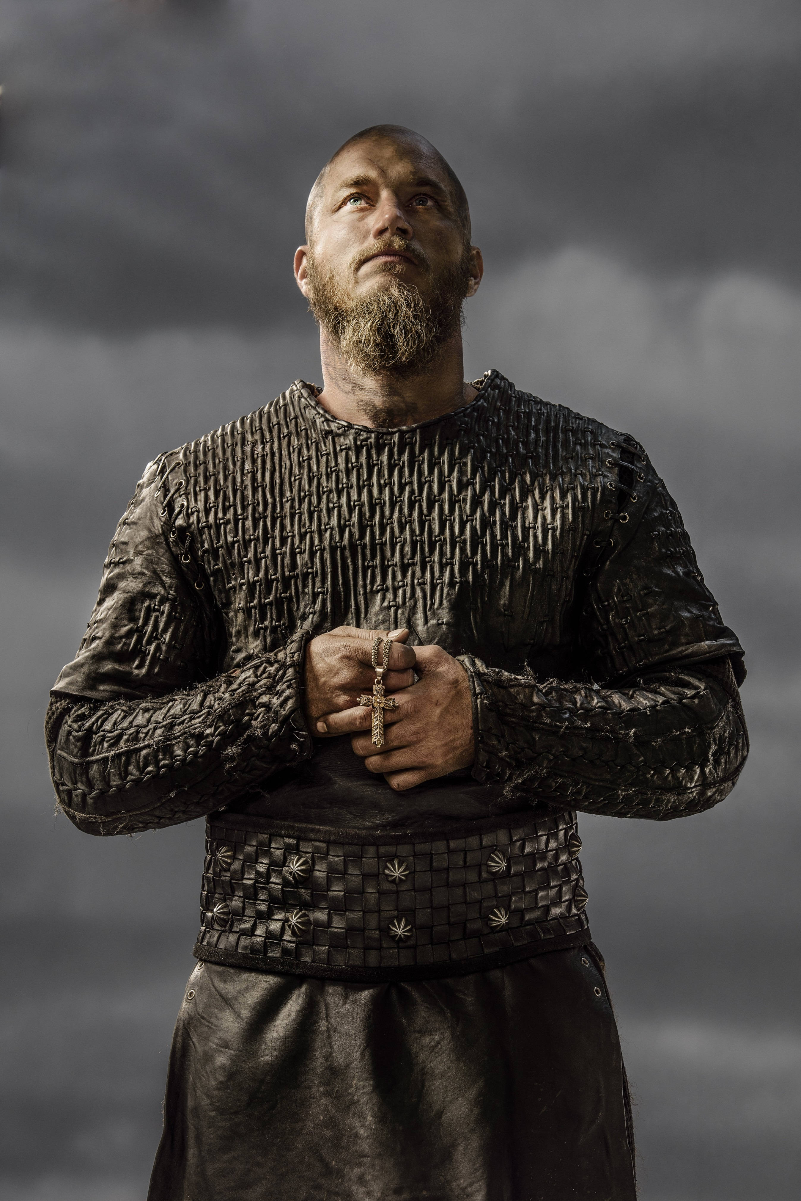Vikings 桌布 壁紙 — 一位留鬍鬚的維京戰士身穿深色鎖子甲，在暴風雨的灰色天空下莊嚴地站立，手持十字架。