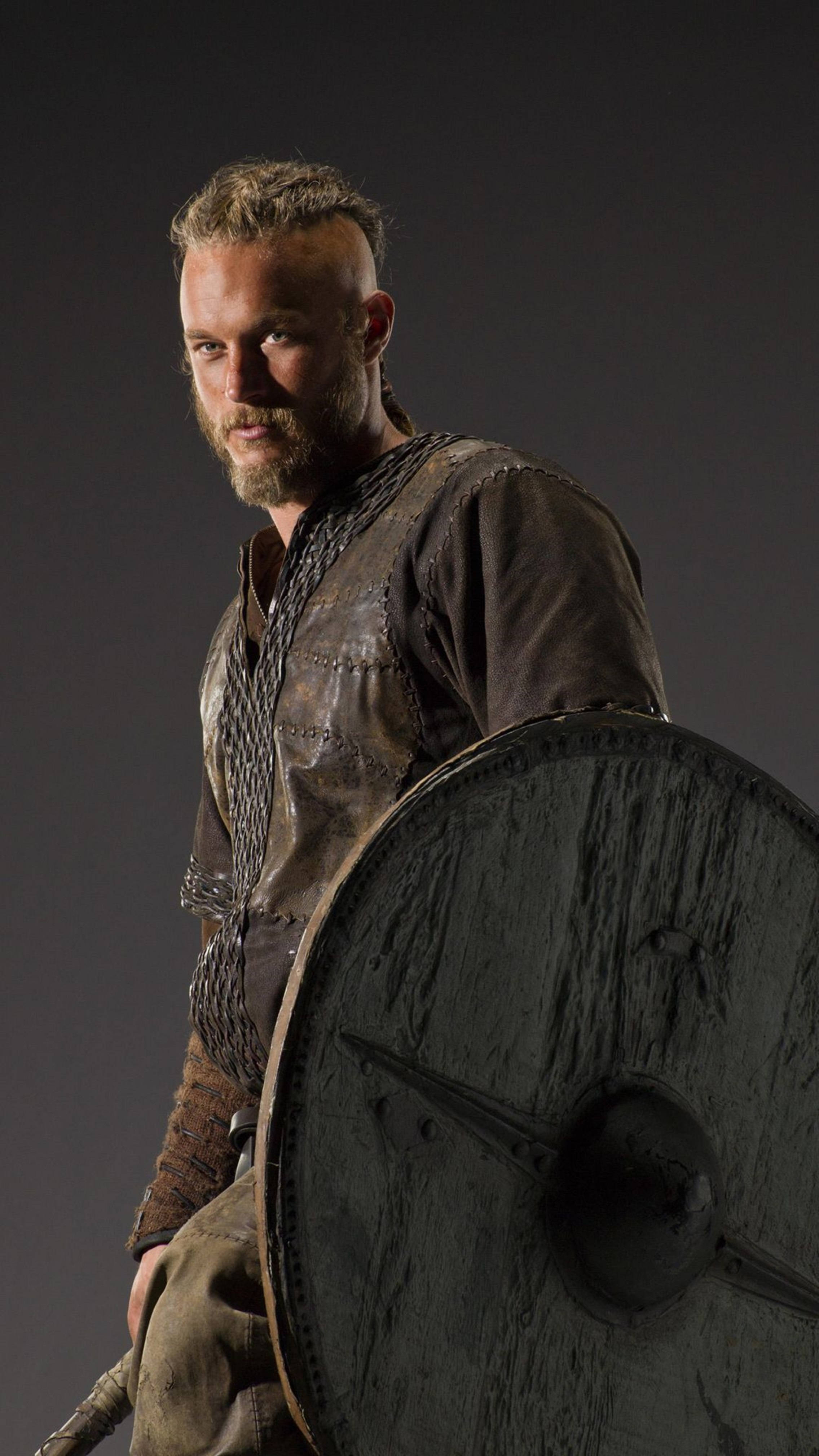 Vikings 桌布 壁紙 — 一位留鬍鬚的維京戰士身穿皮革盔甲，在深灰色工作室背景前手持一個大圓盾。