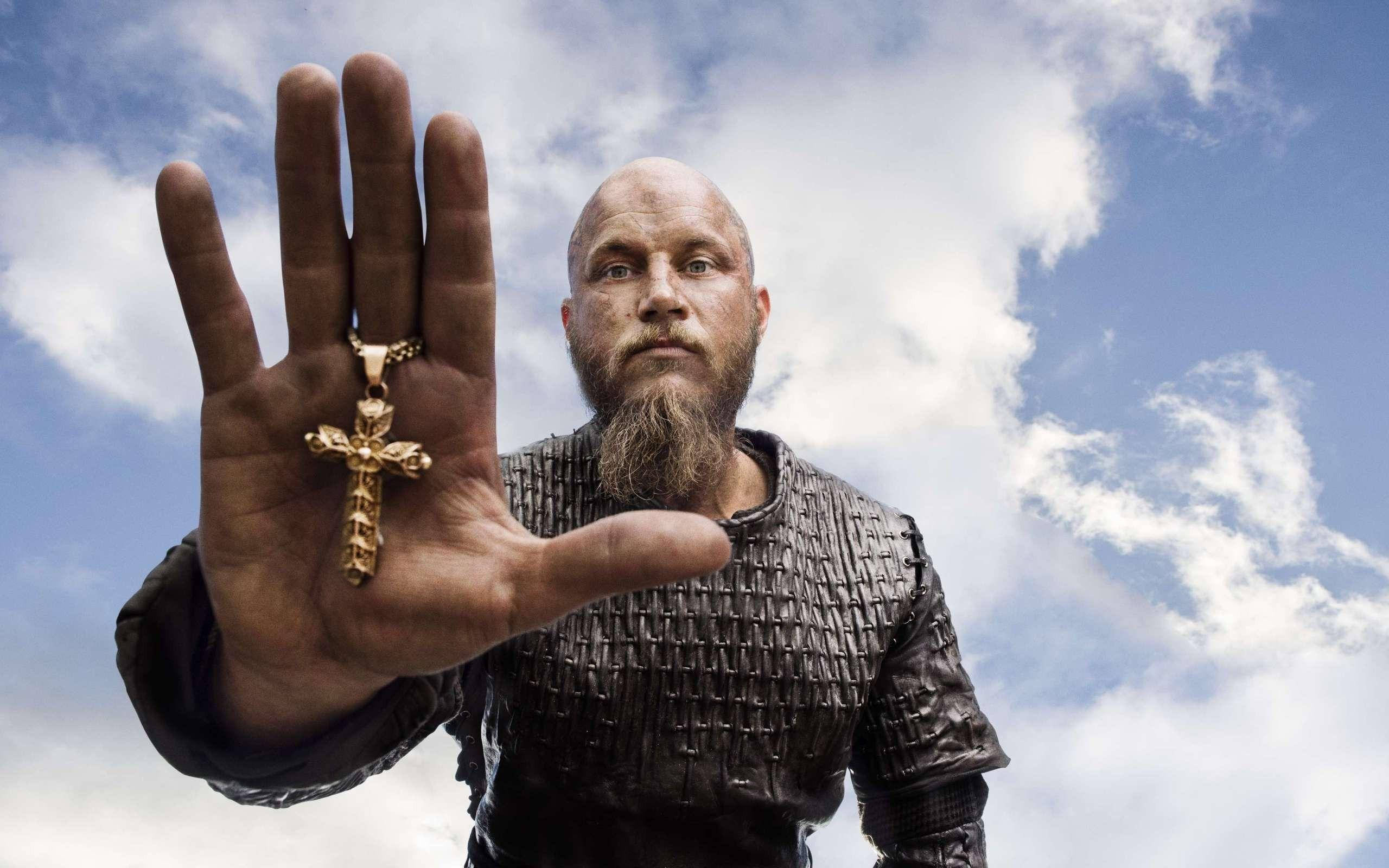 Vikings 桌布 壁紙 — 一位留鬍鬚的維京戰士身穿鎖子甲，舉起一隻手，手上掛著金色十字架吊墜，背景是多雲的藍天。