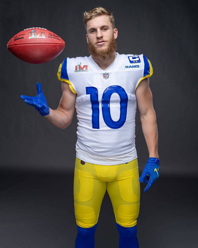 Cooper Kupp 桌布 壁紙 — 一位留鬍鬚的美式足球員，身穿白色、藍色和黃色的公羊隊球衣，在深色攝影棚背景前，用戴手套的手平衡一個紅色美式足球。