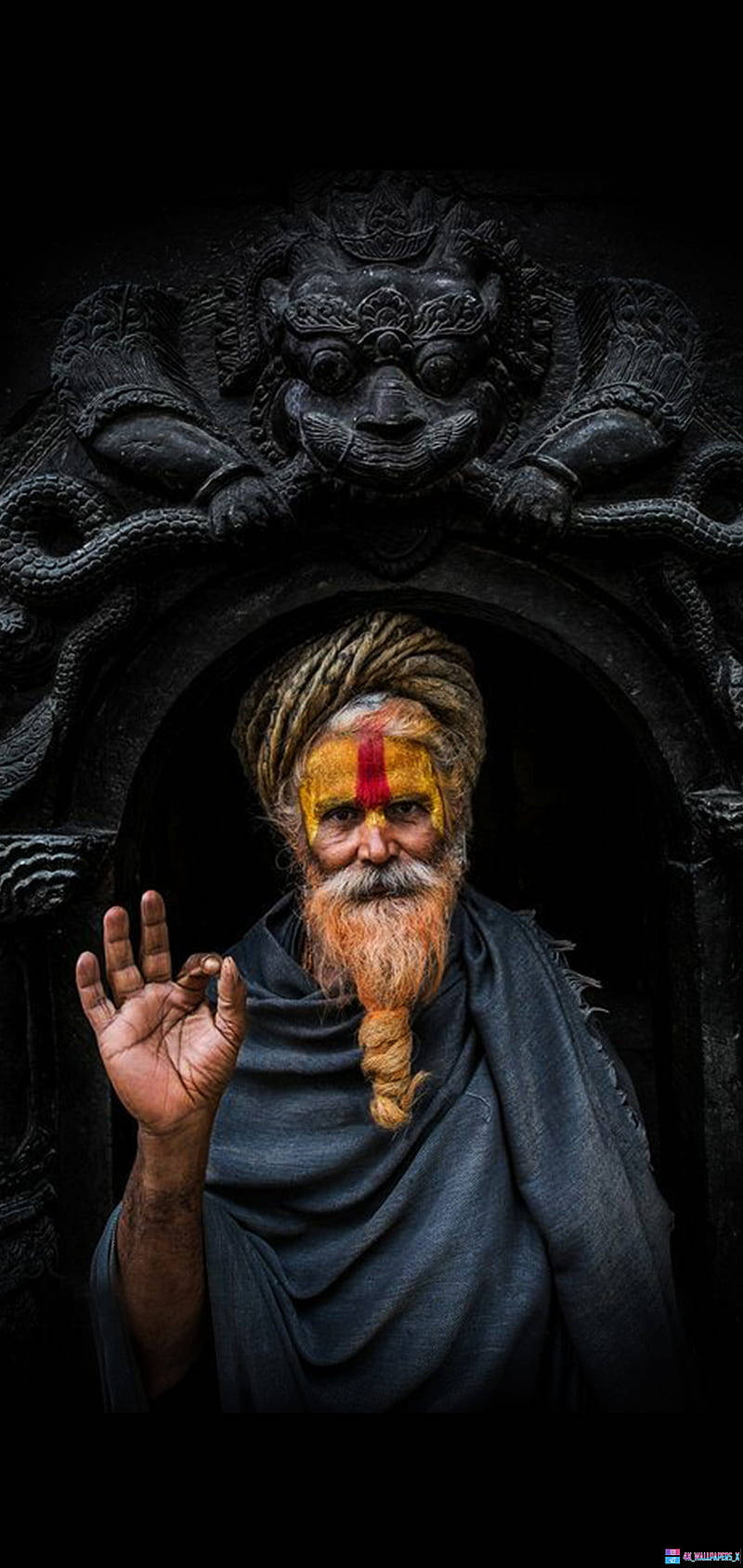 Aghori 桌布 壁紙 — 一位留鬍鬚的聖人，額頭有紅色蒂拉克，頭戴頭巾，站在黑暗的雕刻石拱門中，做出手勢。