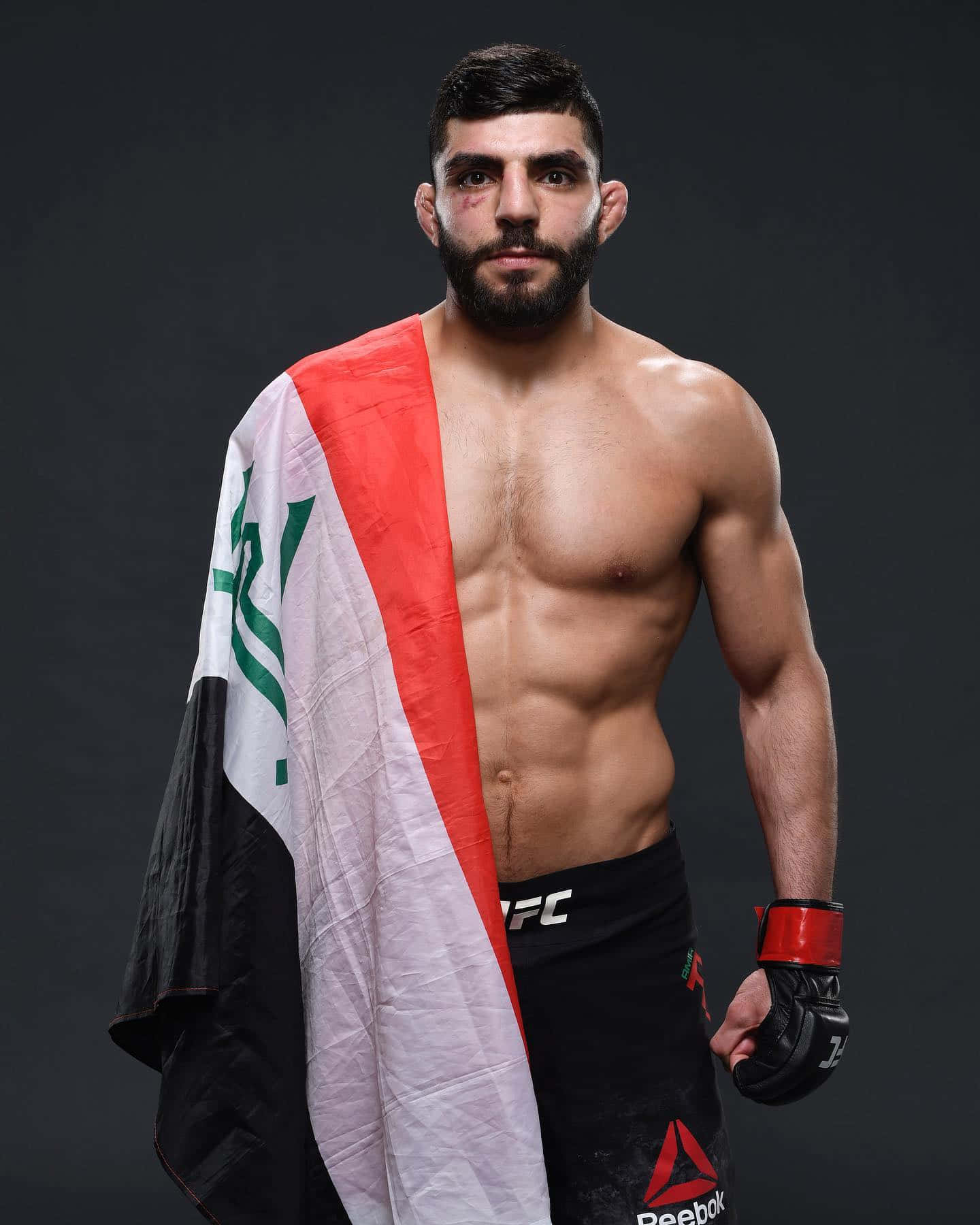 MMA 桌布 壁紙 — 一位留鬍鬚的肌肉發達UFC拳手自信地披著伊拉克國旗，站在深色攝影棚背景前。