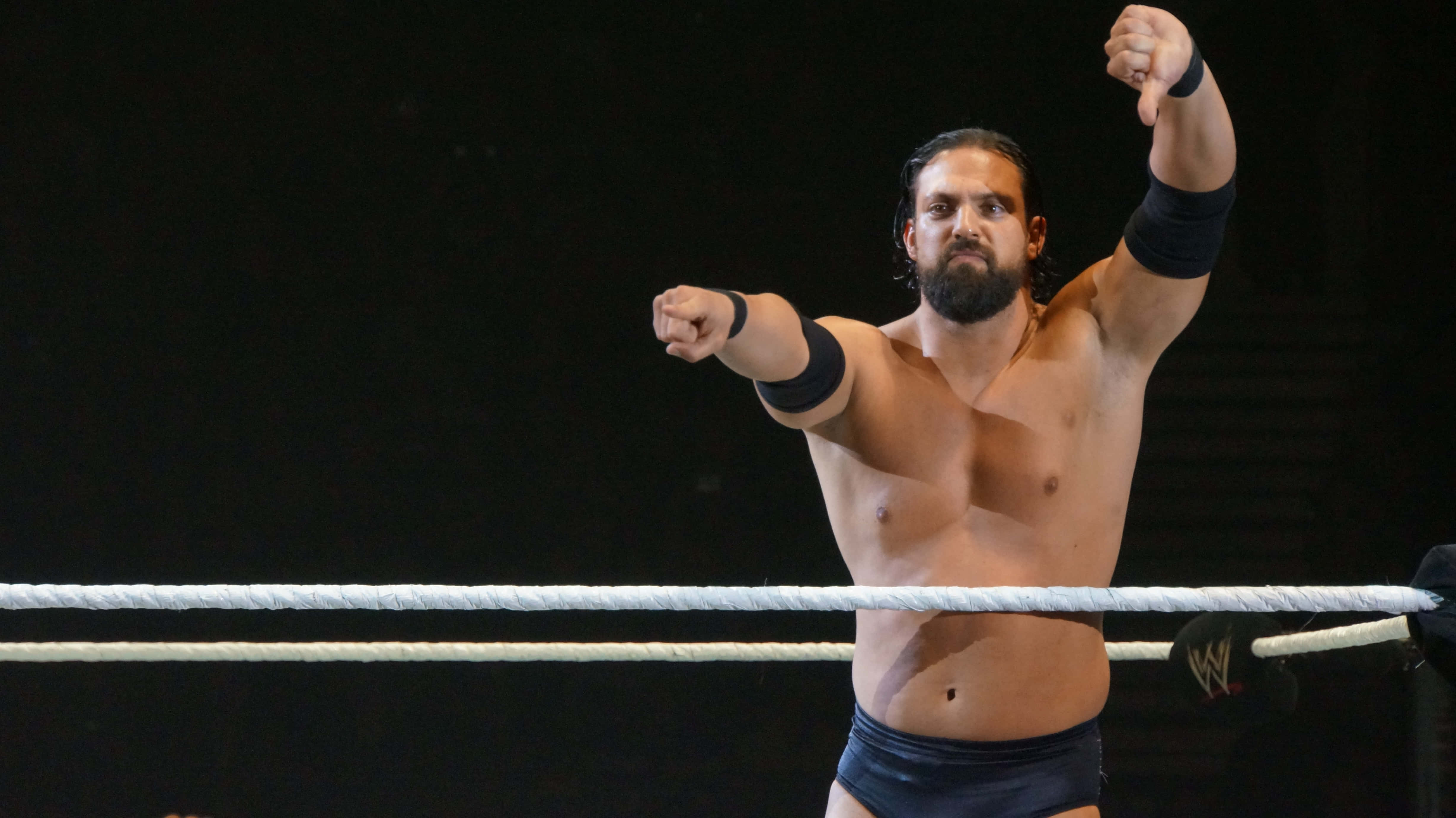 WWE 桌布 壁紙 — 一位留鬍鬚的肌肉發達摔角手在明亮燈光下的摔角場內彎曲手臂並向前指。