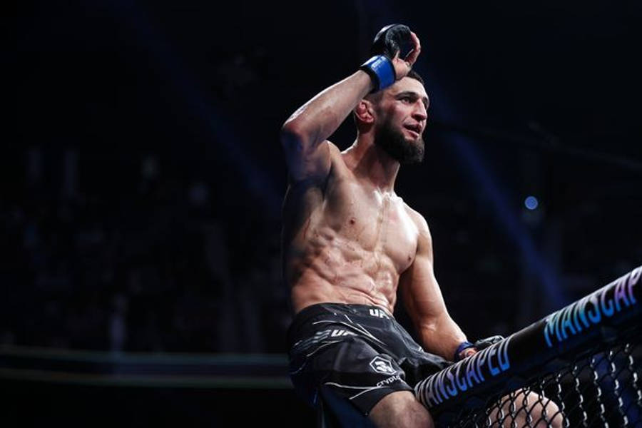 MMA 桌布 壁紙 — 一位留鬍鬚的肌肉發達鬥士坐在昏暗競技場的籠網上，比賽後舉起戴手套的手慶祝勝利。