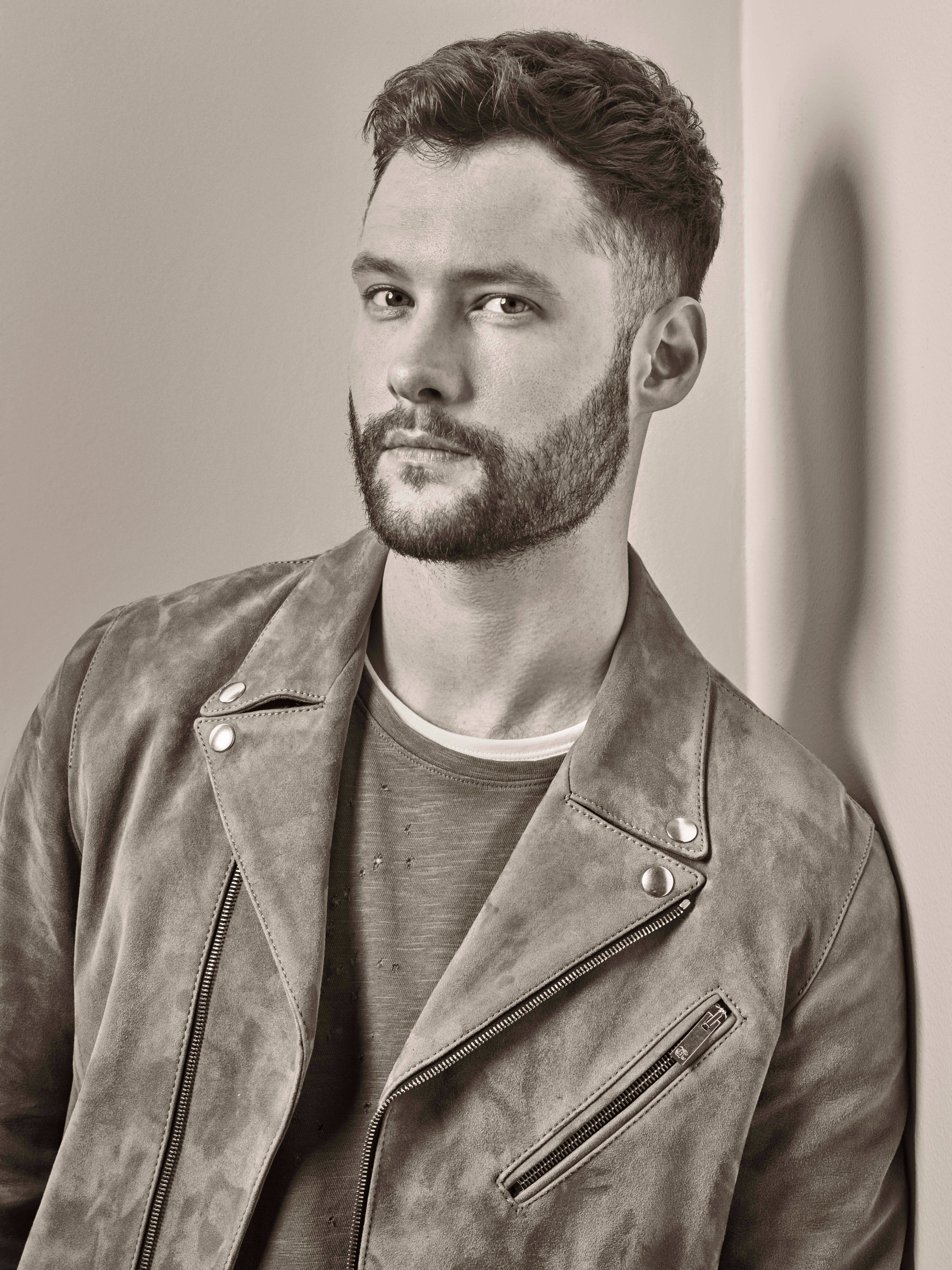 Calum Scott 桌布 壁紙 — 一位留鬍鬚的英俊男士，穿著質感皮革外套，靠著素色牆壁，呈現憂鬱的黑白肖像。