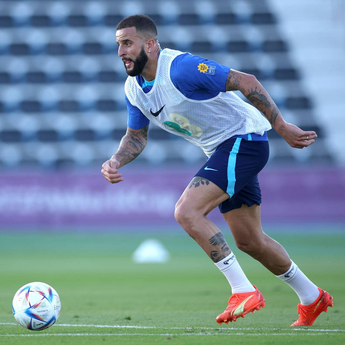 Kyle Walker 桌布 壁紙 — 一位留鬍鬚的足球員身穿藍白裝備，在綠色球場上運球，身後是空無一人的球場座位。